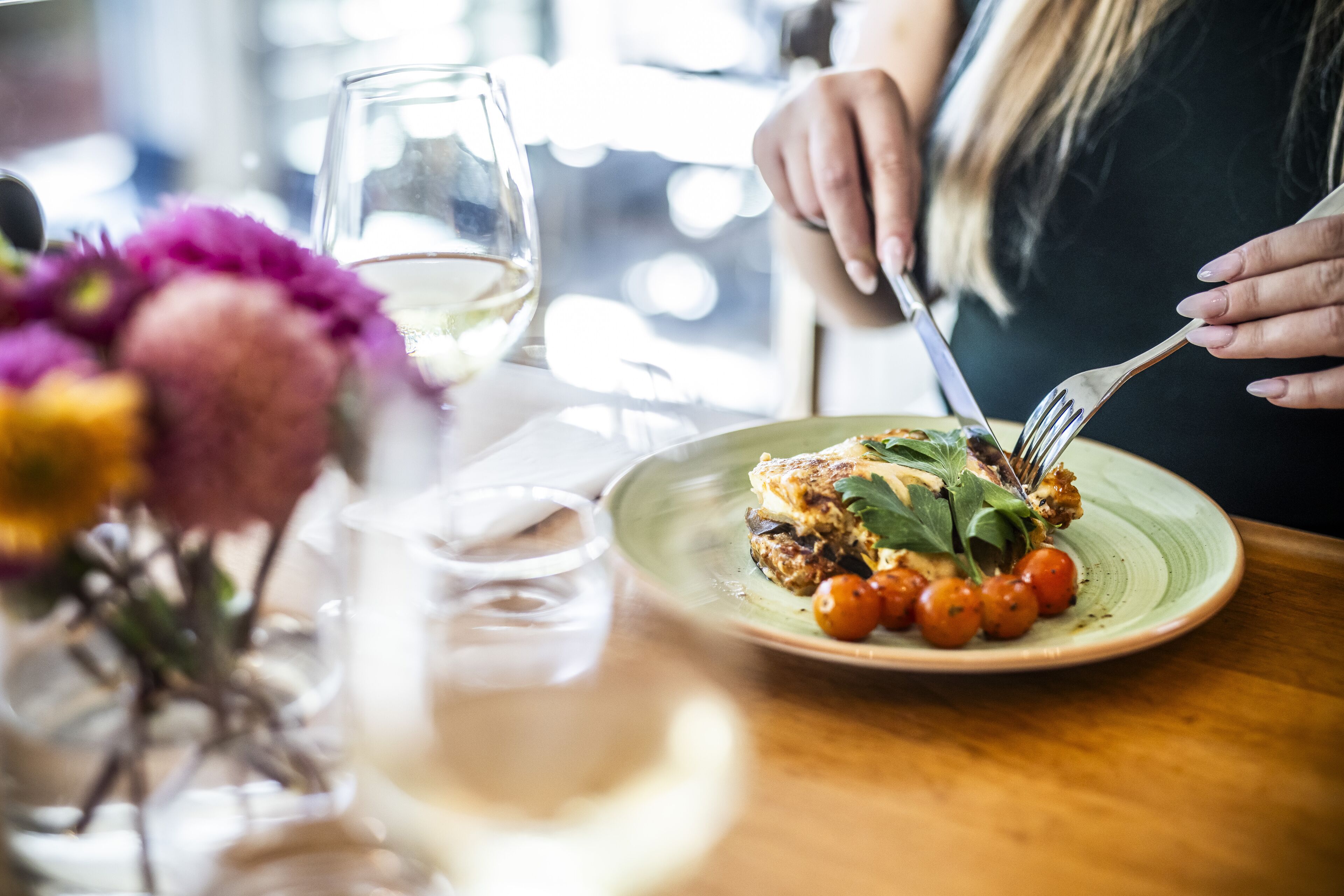 Une table à manger avec une assiette de nourriture, un verre de vin blanc et un vase rempli de fleurs.