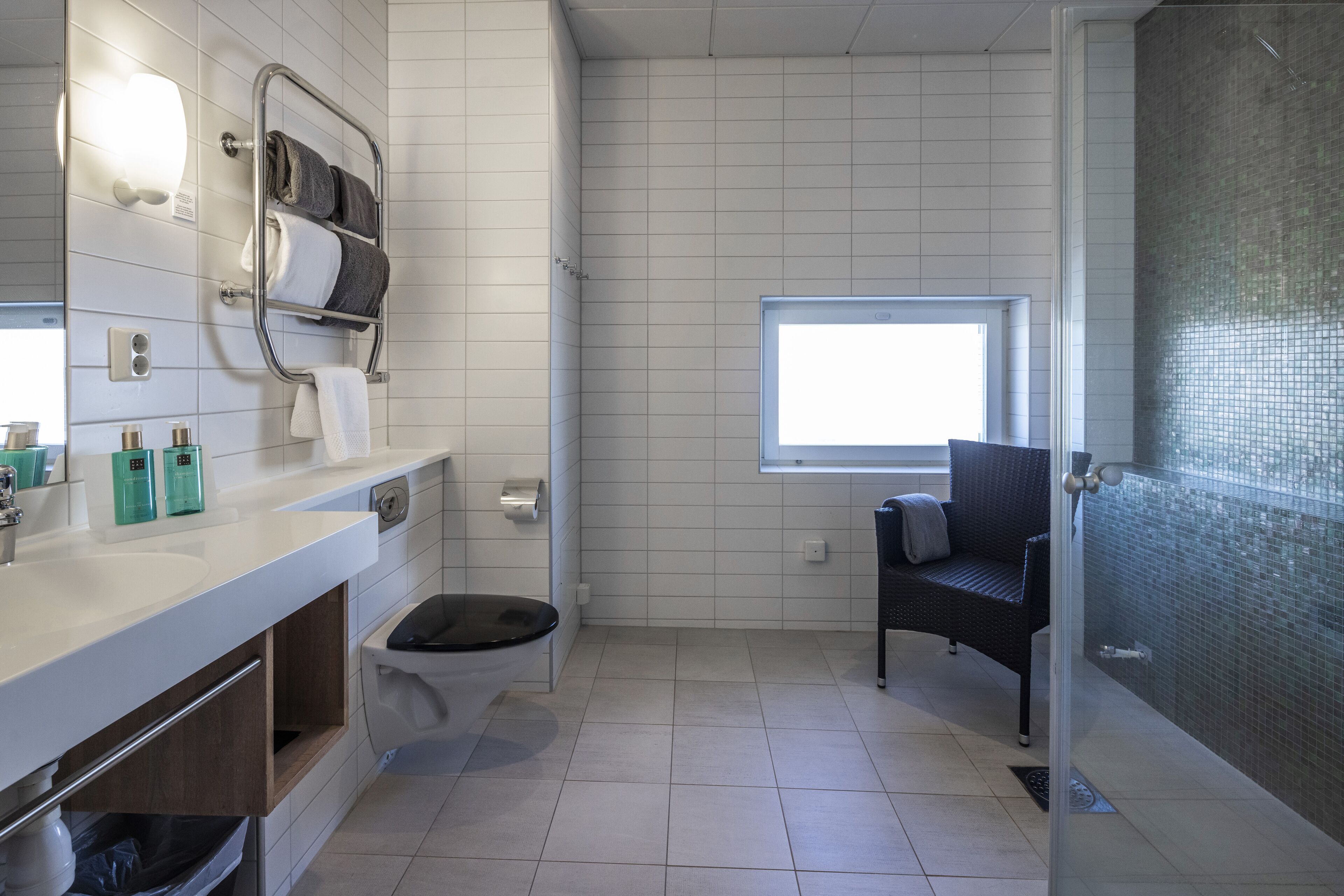 A modern bathroom with a white sink, toilet, and shower area. There is a towel rack with towels, a window providing natural light, and a chair near the shower.