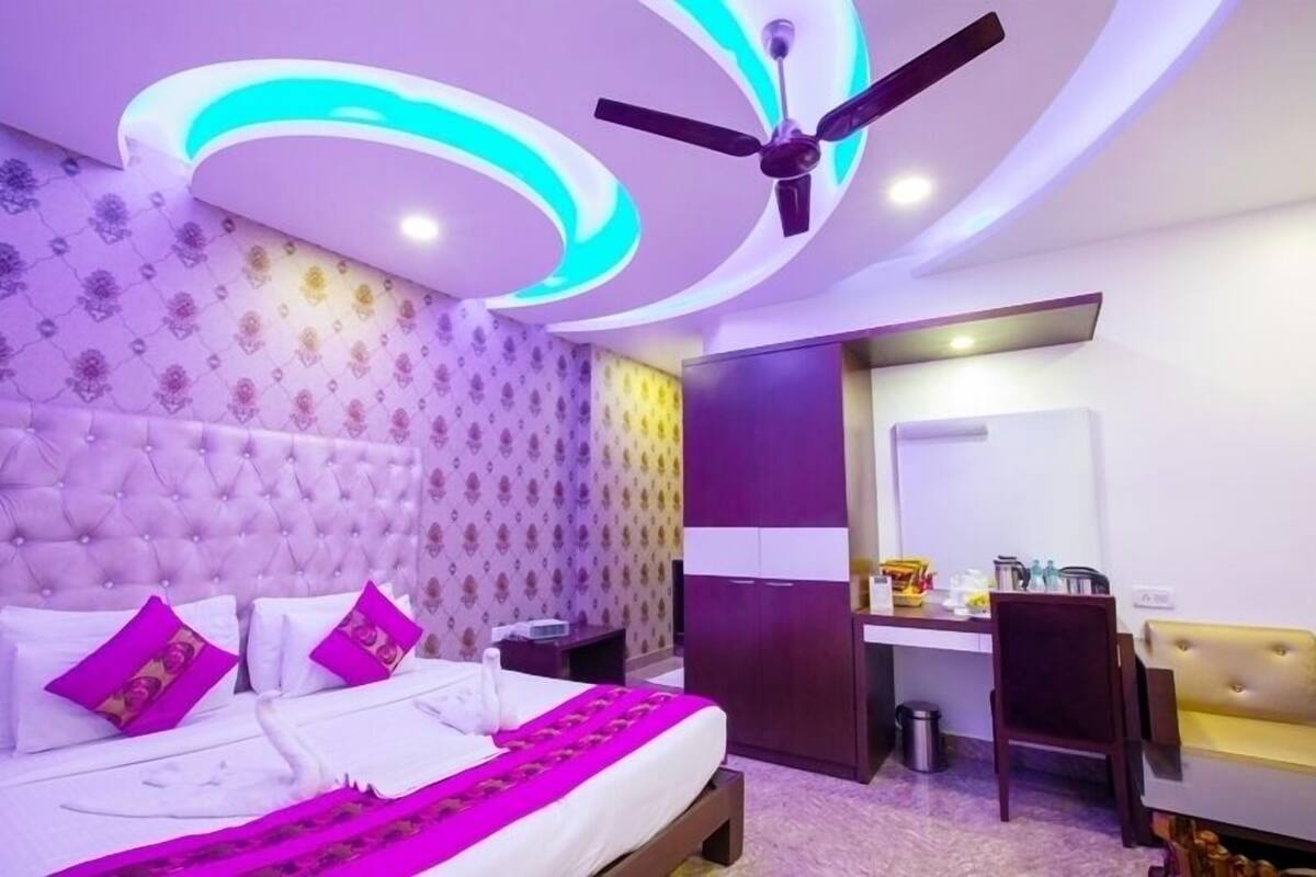Un dormitorio con cabecera capitonada, cama con ropa de cama blanca y detalles morados, un escritorio con silla y un ventilador de techo.