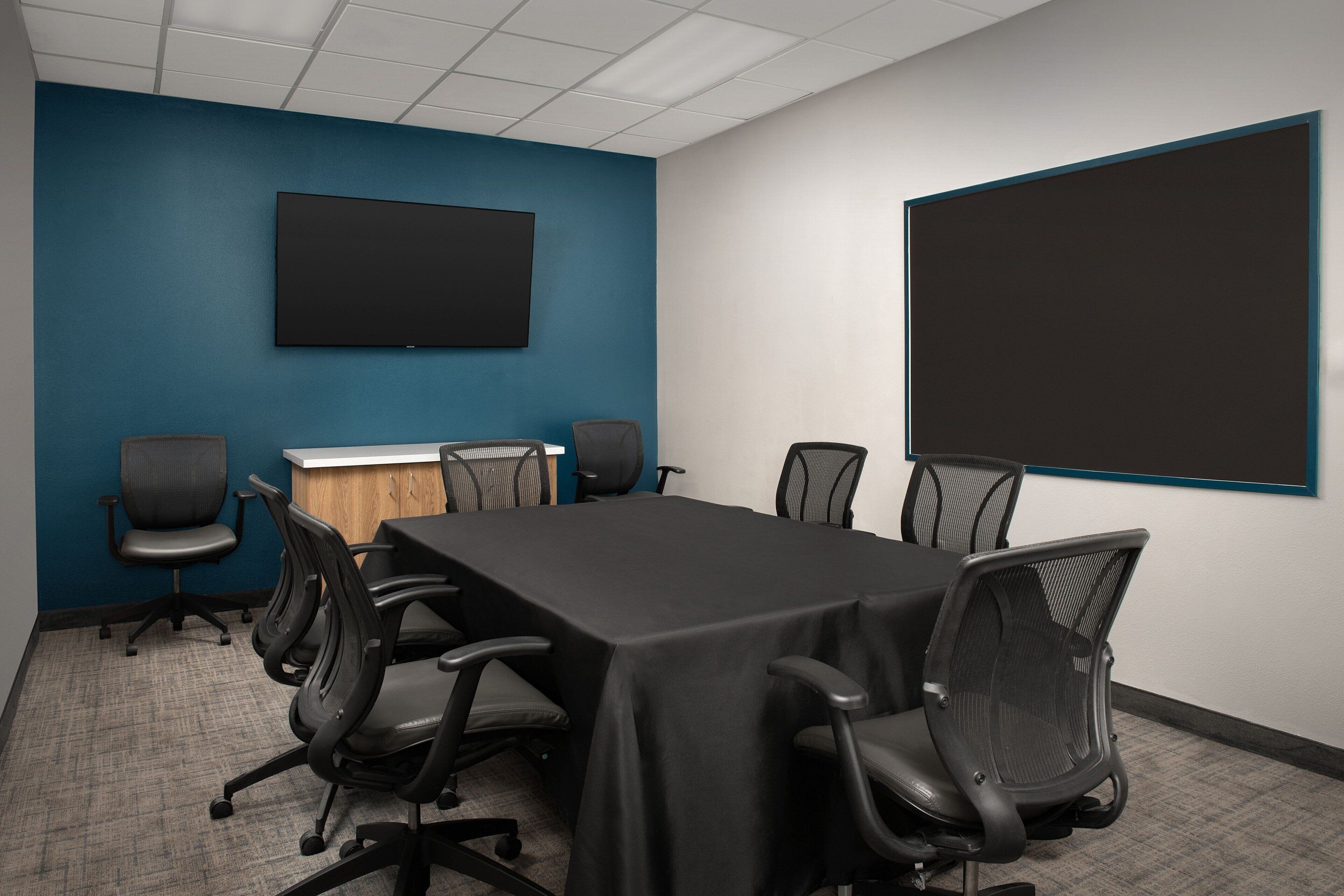 A conference room with a long table, six chairs, a TV on the wall, and a large screen.