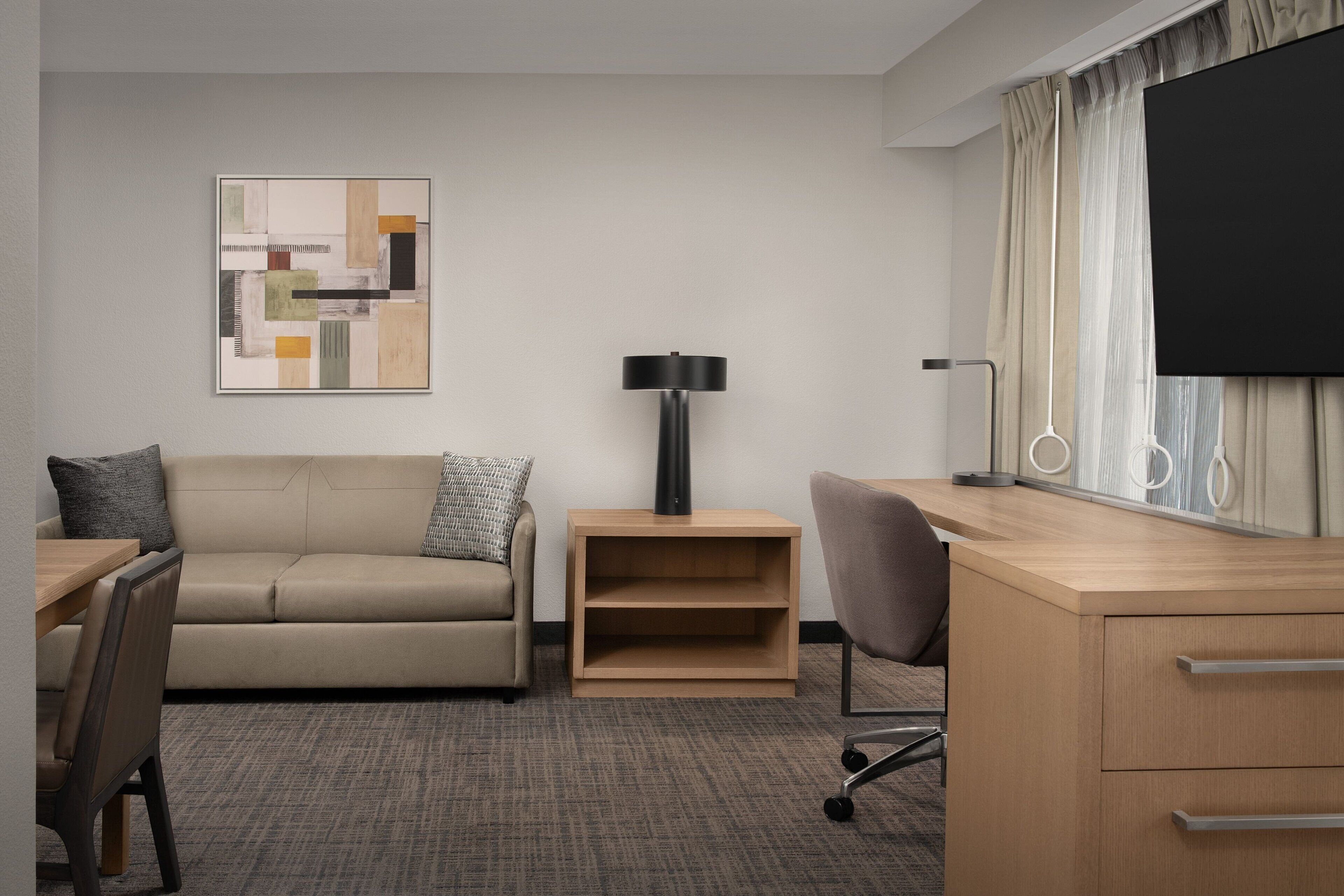 A hotel room with a sofa, a desk, a chair, a lamp, a TV, and a wall art piece.