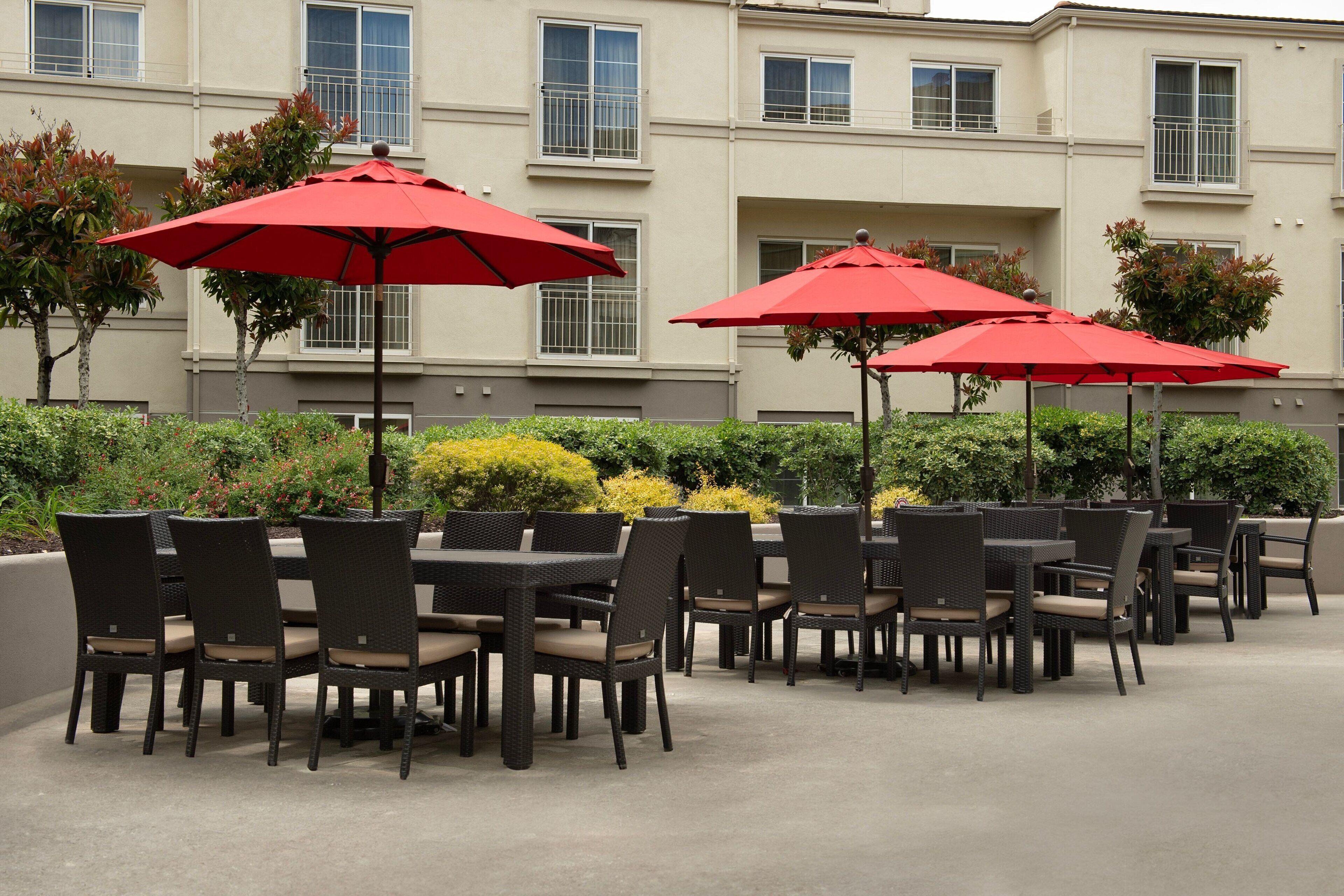 Outdoor patio seating with tables and chairs, red umbrellas, and a building in the background.