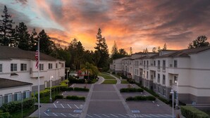 A parking lot with marked spaces, a flagpole with an American flag, and a row of buildings with a central pathway. - Residence Inn Marriott Palo Alto Los Altos (Los Altos)