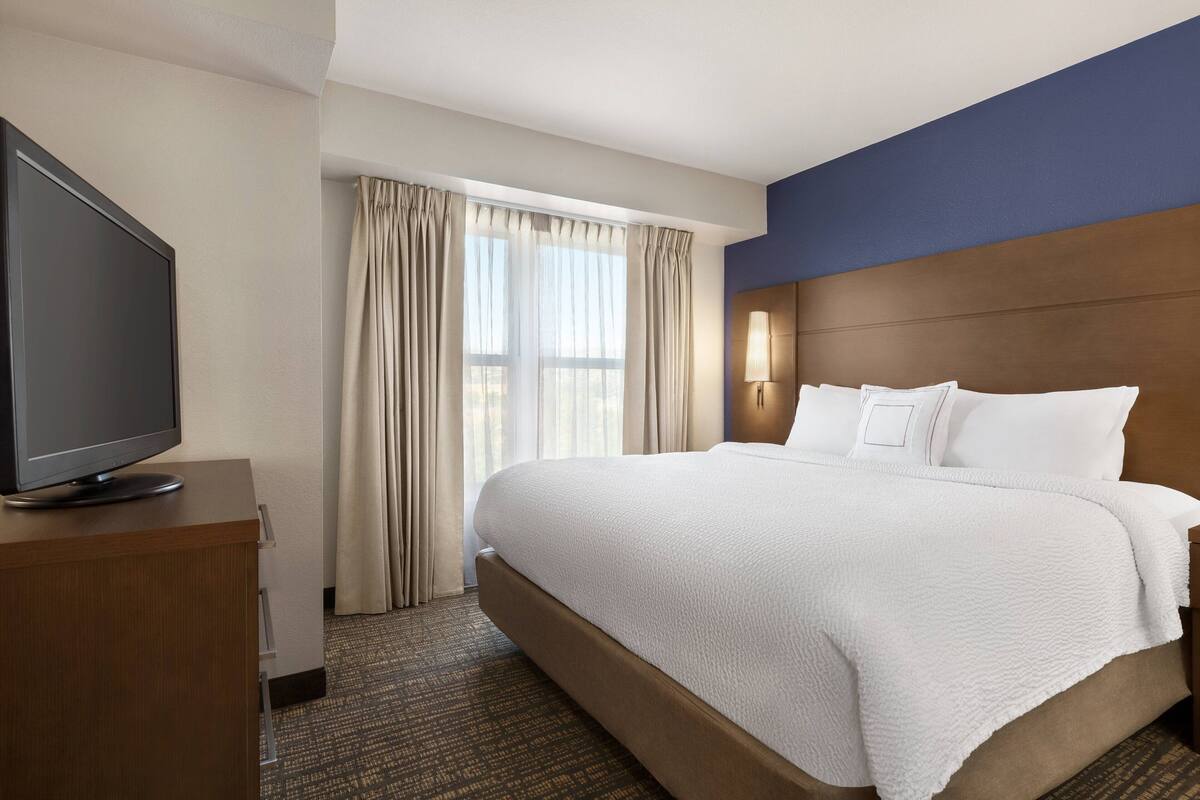 A hotel room with a large bed, a flat-screen TV on a wooden cabinet, and a window with curtains.