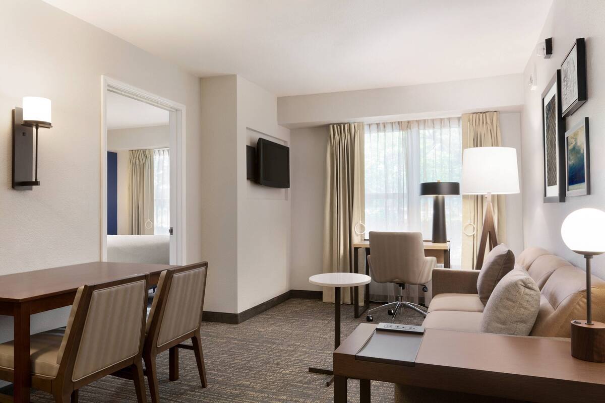 A hotel room with a dining table, chairs, a sofa, a TV, and a lamp.