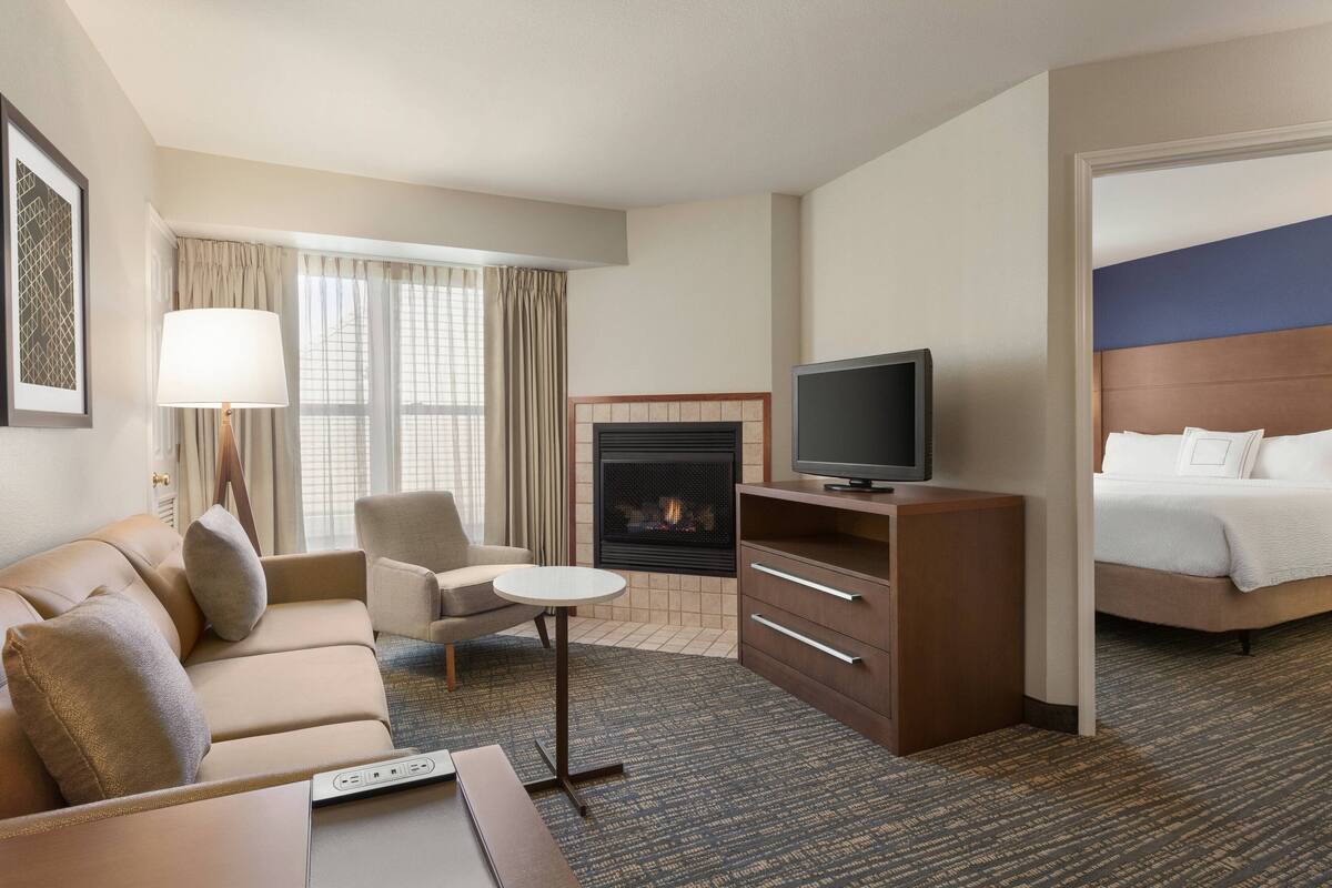 A hotel room with a sofa, armchair, TV, fireplace, and a bed.