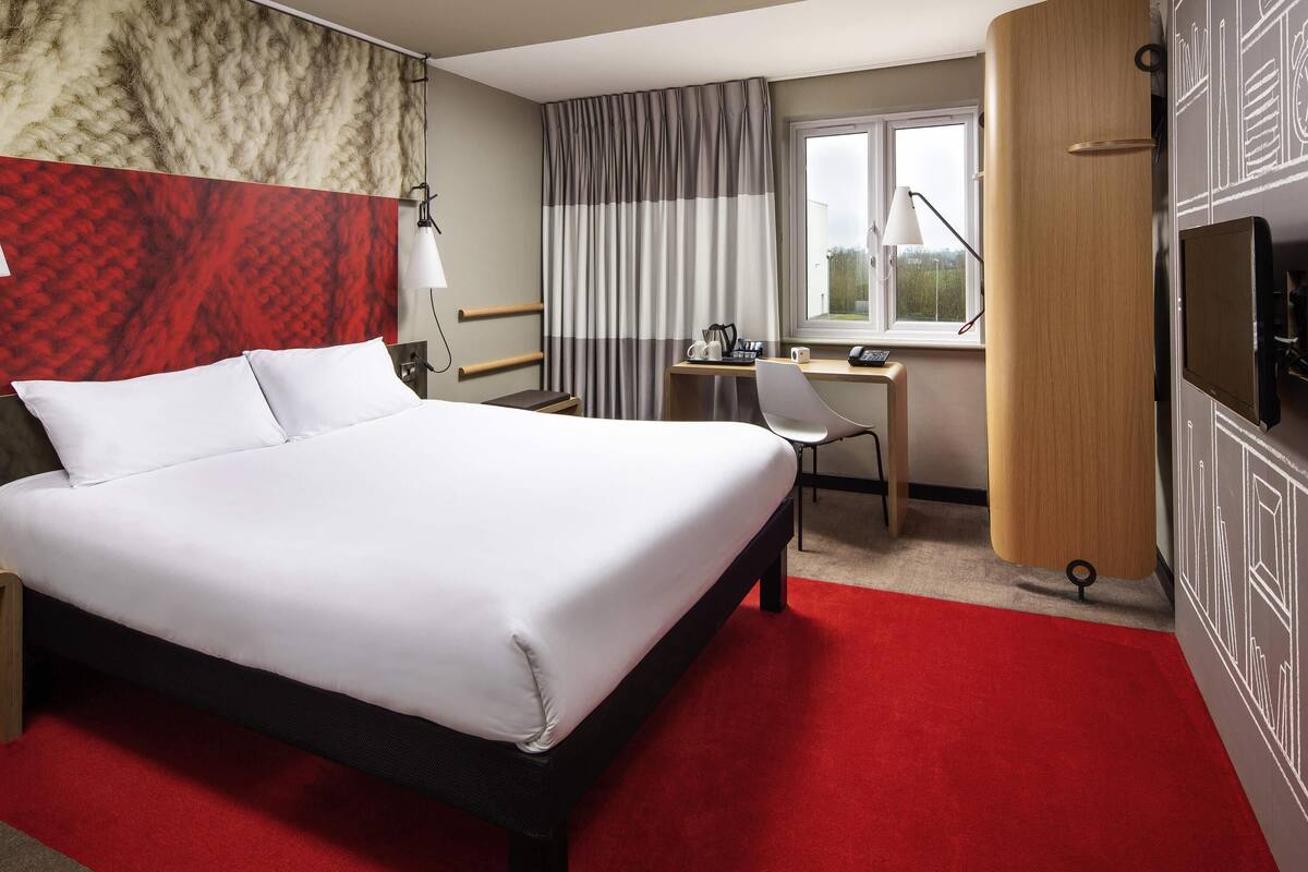 A hotel room with a large bed, a desk, and a chair. There is a window with curtains, a lamp, and a TV on the wall.