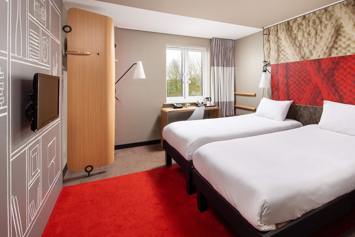 A hotel room with two beds, a desk with a chair, a TV, a wardrobe, and a window with curtains.