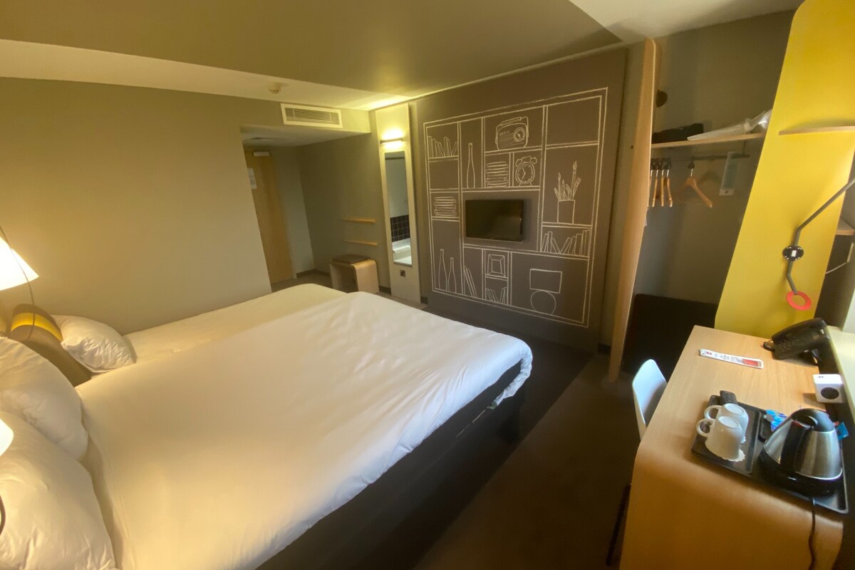 A hotel room with a bed, a desk with a coffee maker, a telephone, and a wall with a built-in TV and storage compartments.