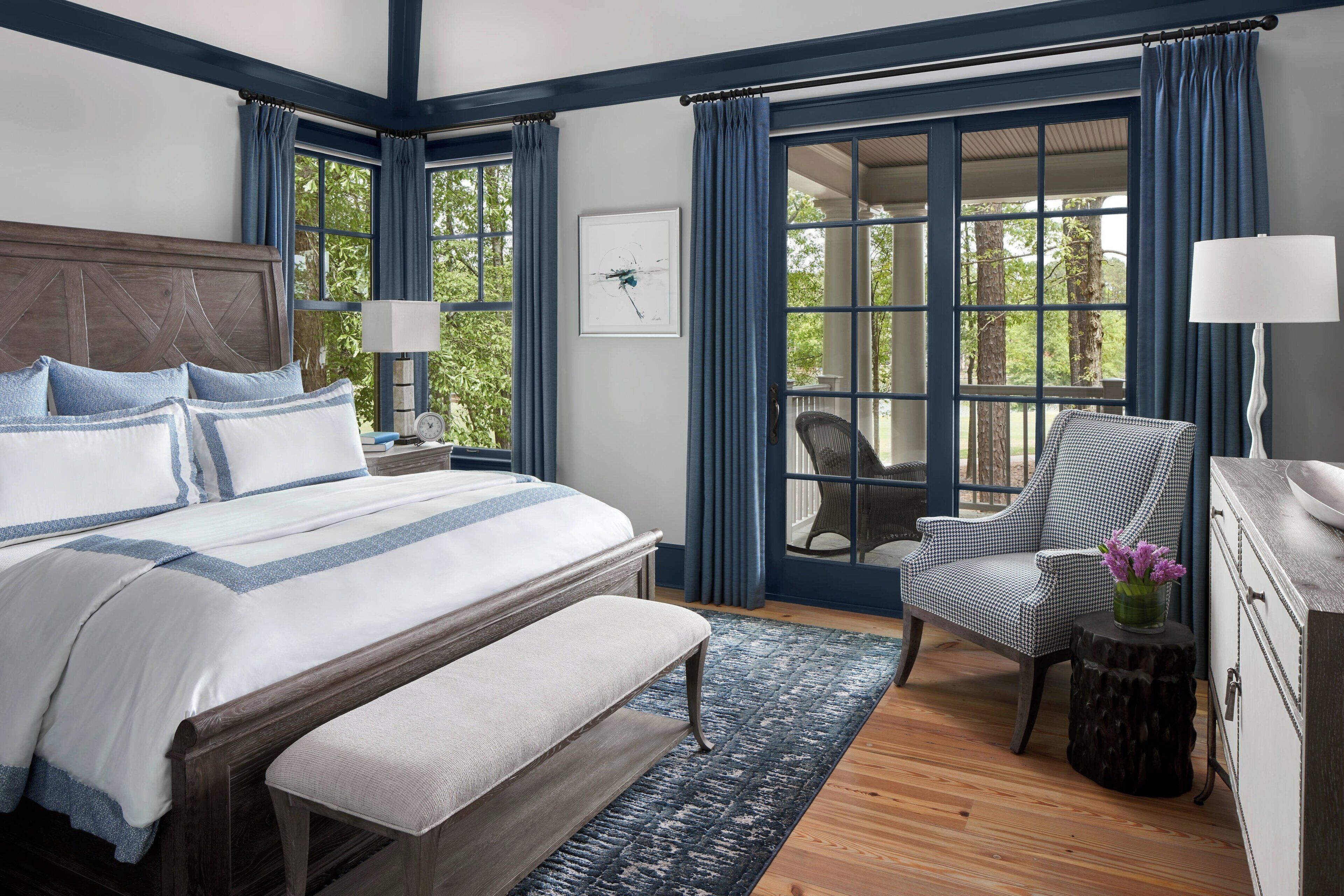 A bedroom with a large bed, a bench, a chair, a small table with a lamp, and a view through a large window.