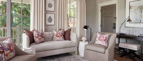 Un salón con un sofá color crema, dos sillones, una mesita auxiliar y un escritorio con una lámpara. Hay cortinas en las ventanas y cuadros en las paredes.