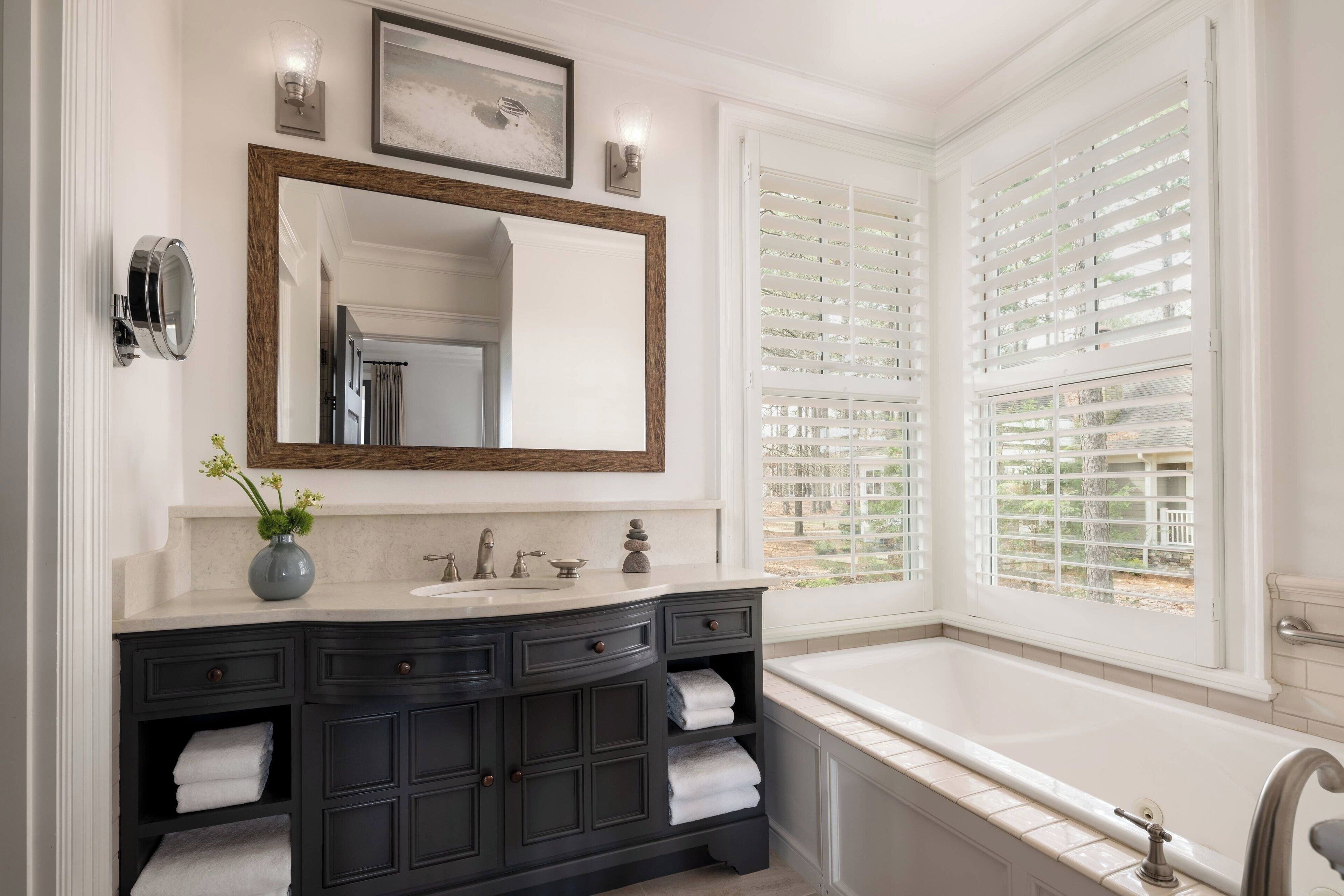 A bathroom with a large mirror, dark wooden vanity, white bathtub, and a window with blinds.