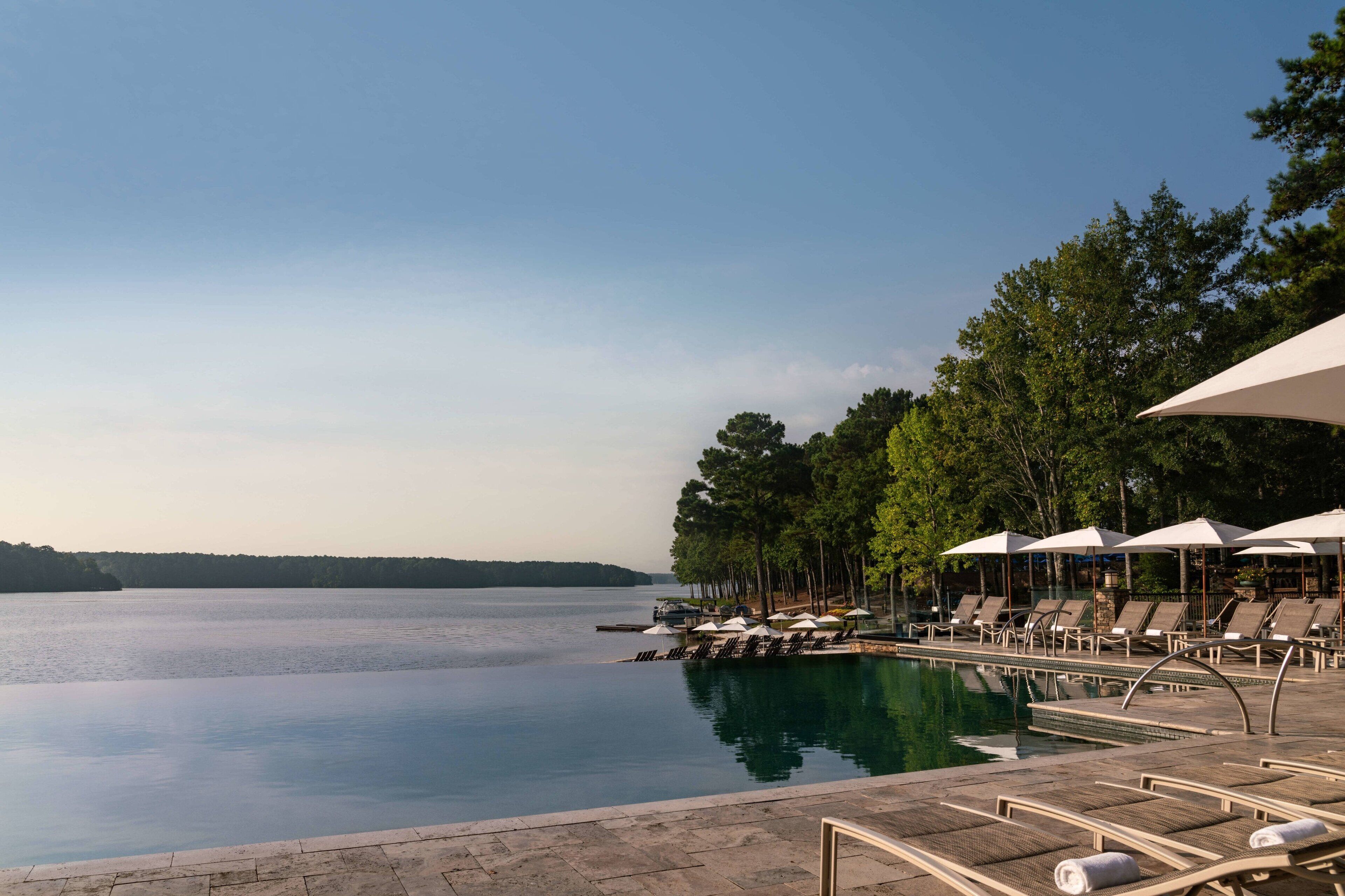 A serene pool area with lounge chairs and umbrellas, surrounded by trees and a calm lake.