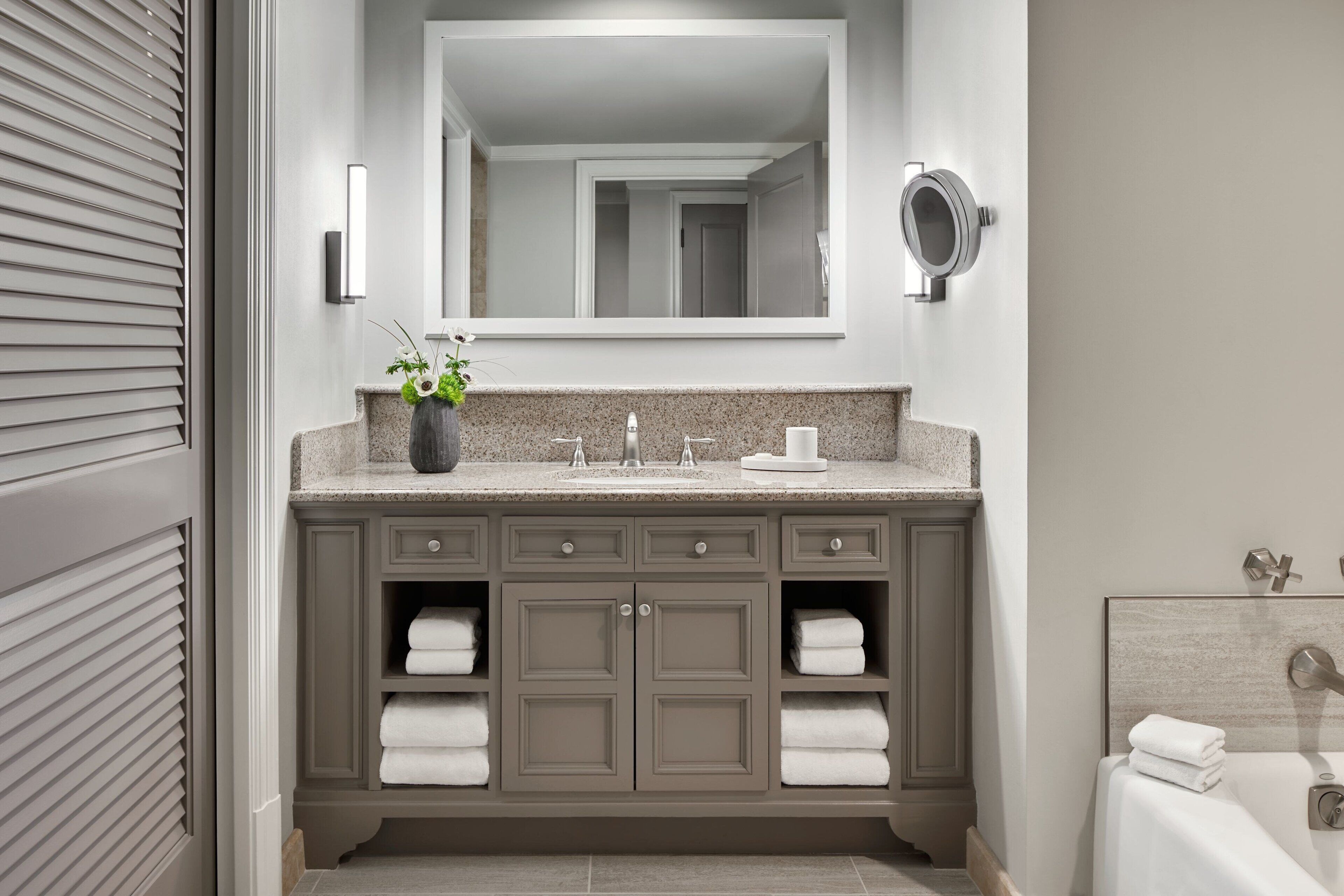 A modern bathroom with a large mirror, a granite countertop, and a built-in sink. There are two towel racks and a small plant on the vanity.