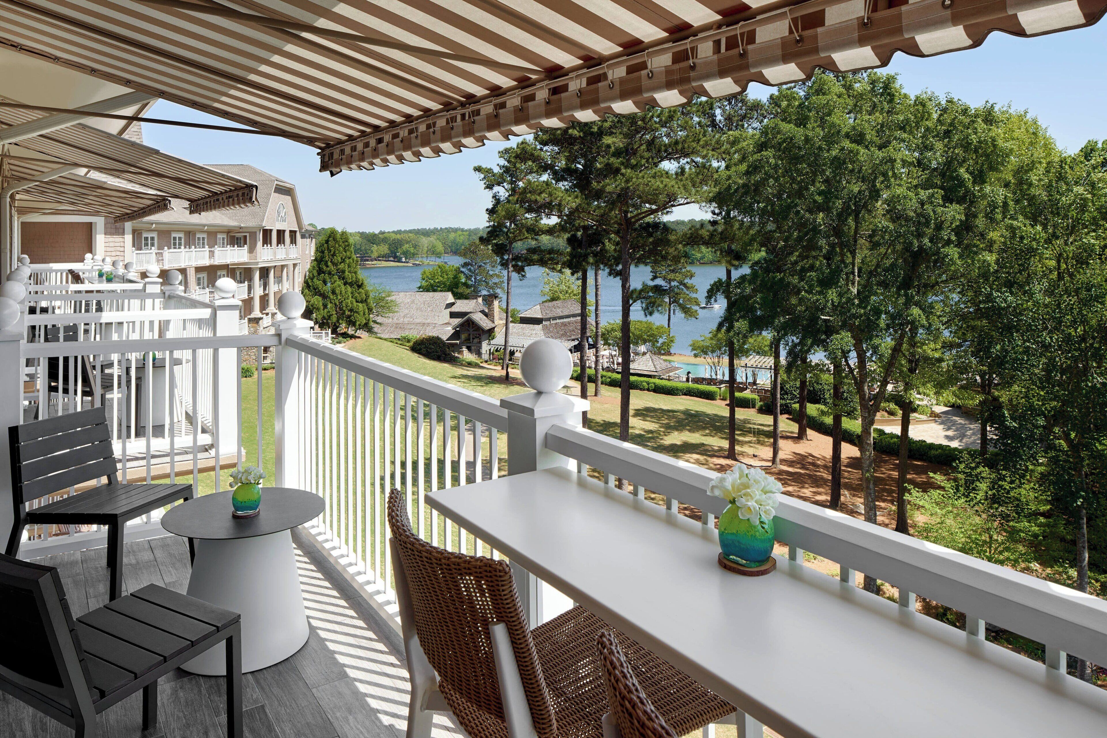 A balcony with a white railing, a table, and two chairs. There is a view of a lake and trees in the background.