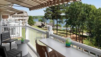 A balcony with a white railing, a table, and two chairs. There is a view of a lake and trees in the background.
