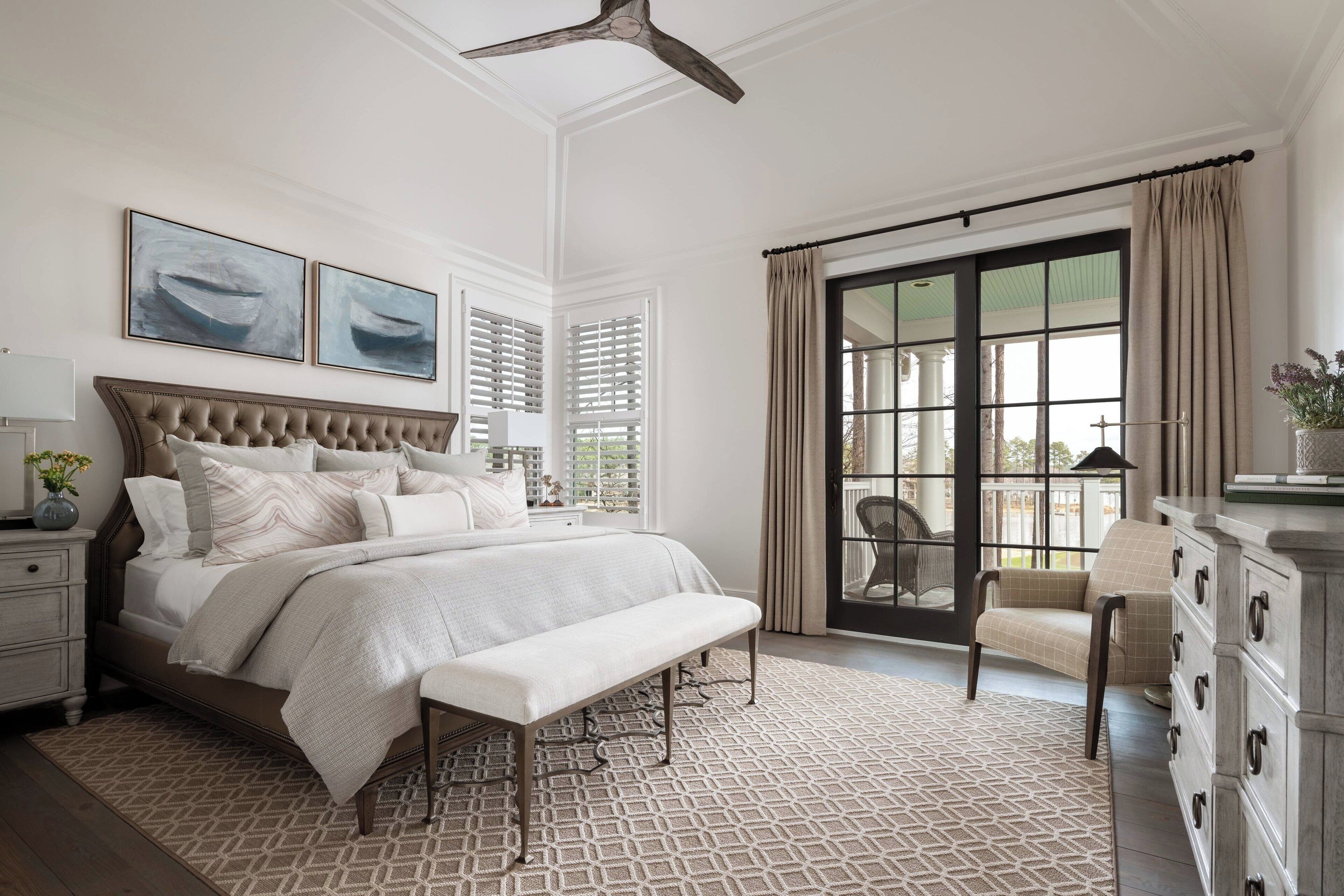 A bedroom with a large bed, a bench, a dresser, and a chair. There are two windows with shutters and a view of the outside.
