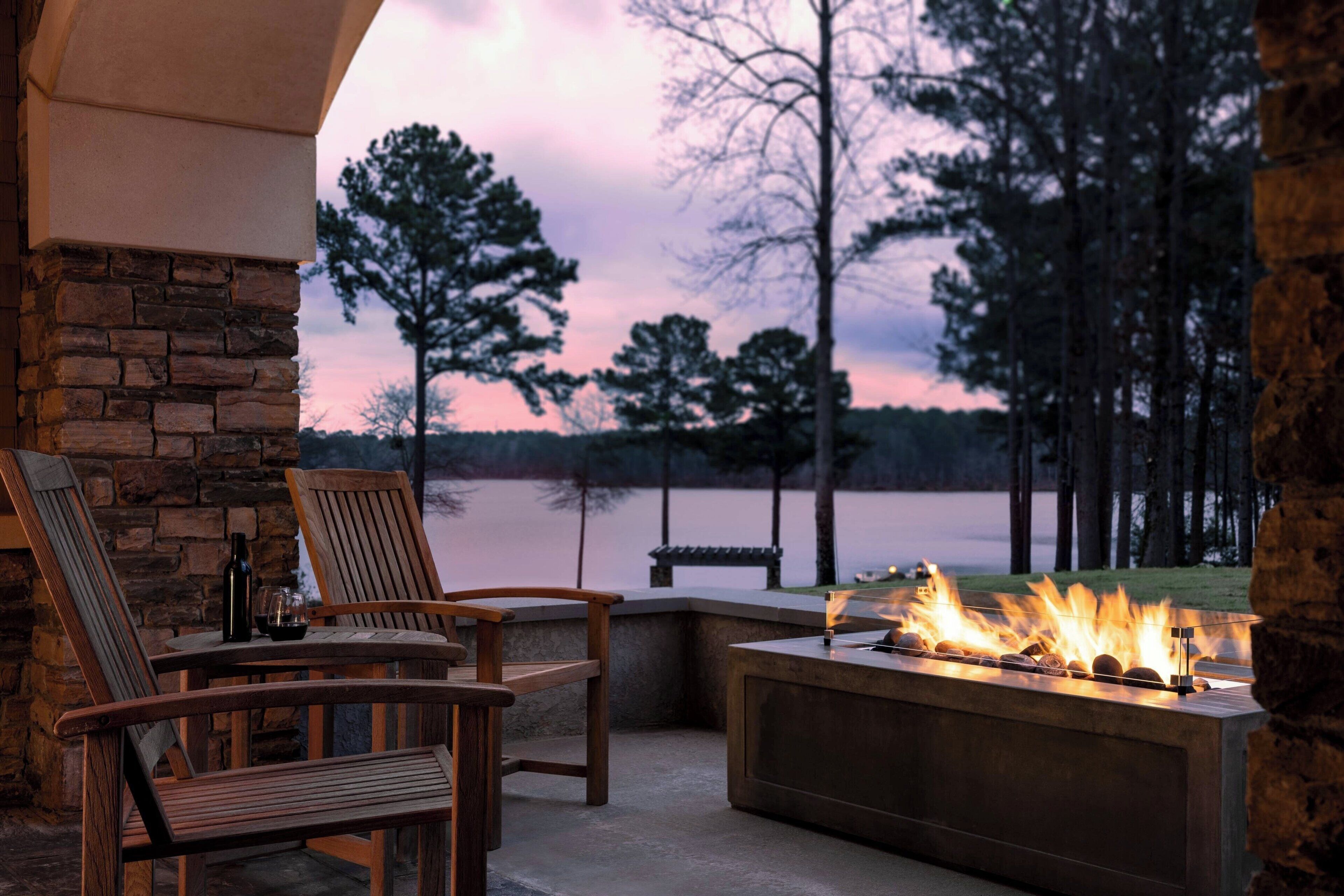A stone wall, wooden chairs, a table with a bottle and glass, and a fire feature with logs.