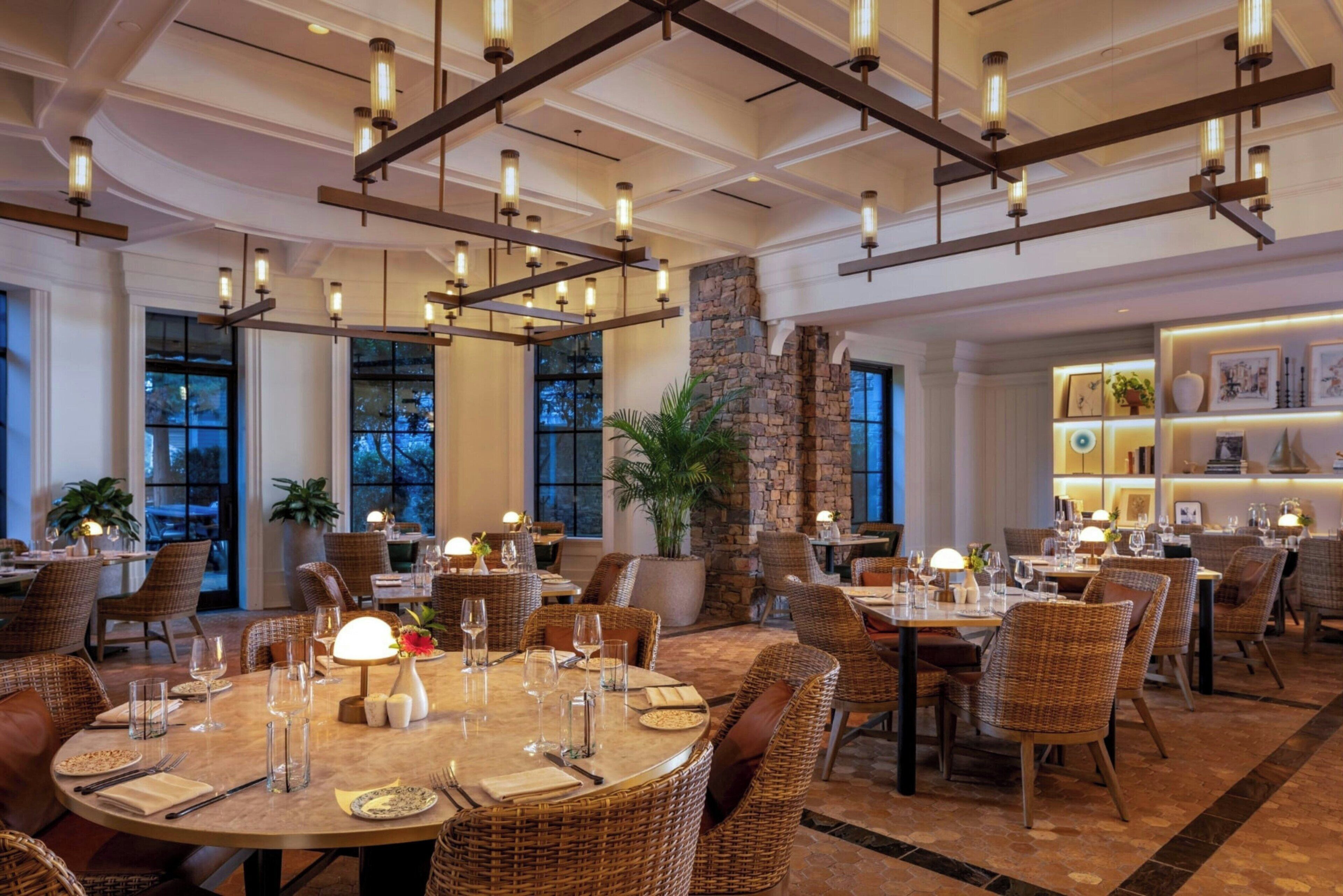 A restaurant with round tables, wicker chairs, and pendant lighting.