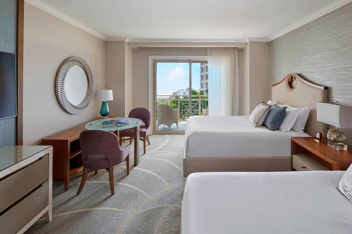A hotel room with two beds, a desk, a chair, a round table, and a large window with a view of buildings.