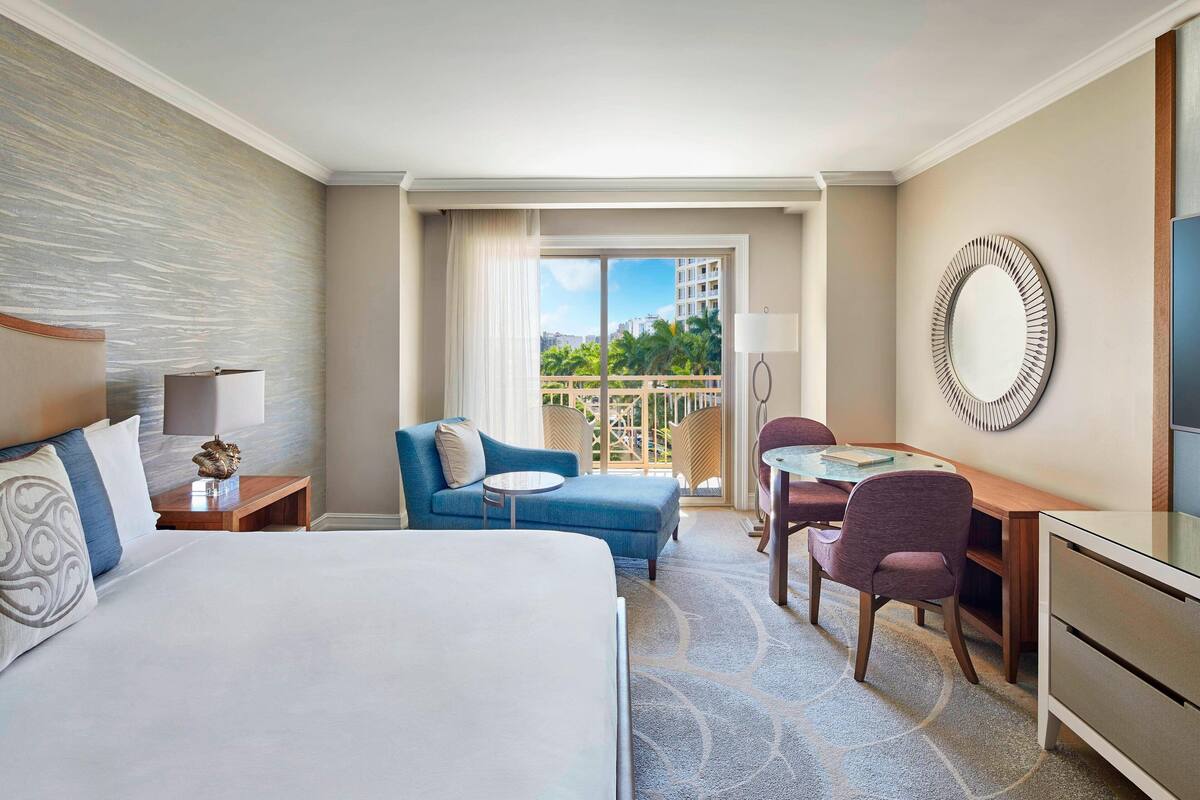 A hotel room with a large bed, a blue sofa, a desk with a chair, a round mirror, and a view of the city through the window.