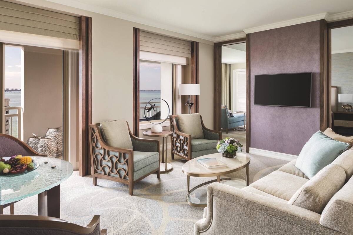 A spacious living room with a large sofa, two armchairs, a coffee table, and a TV mounted on the wall. There is a balcony with a view of the water.