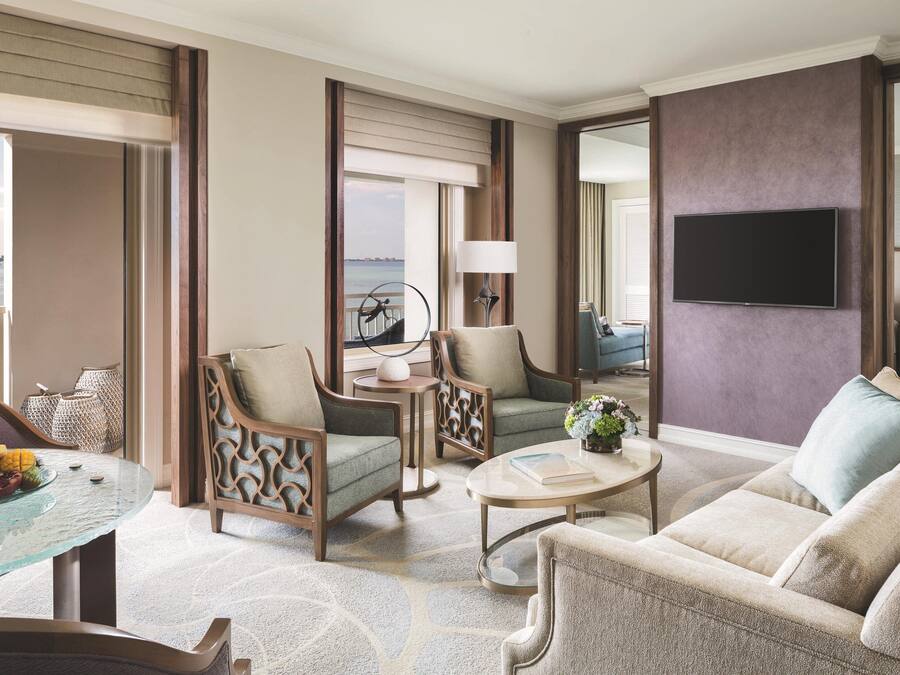 Ein geräumiges Wohnzimmer mit einer großen Couch, zwei Sesseln, einem Couchtisch und einem an der Wand befestigten Fernseher. Es gibt einen Balkon mit Blick aufs Wasser.