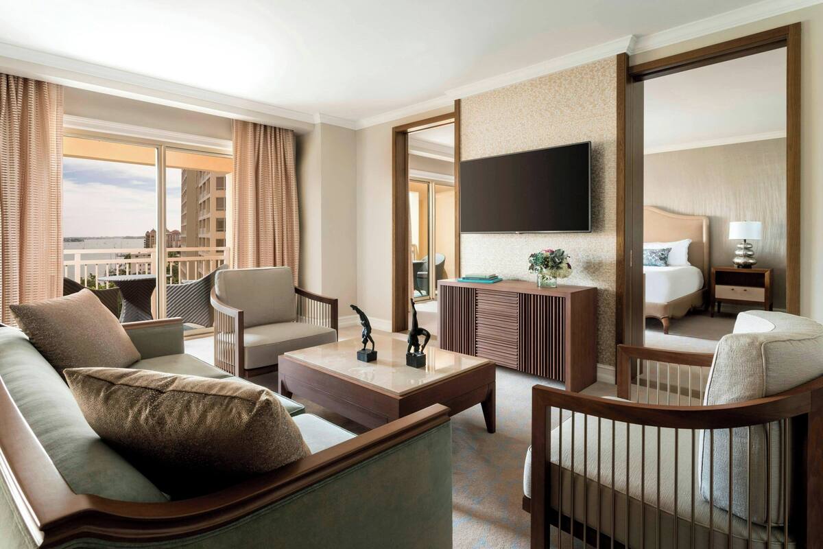 A hotel room with a large bed, a TV mounted on the wall, a seating area with a sofa and chairs, and a balcony with a view of the city.