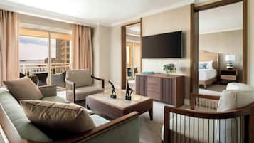 Habitación de hotel con una cama grande, un televisor montado en la pared, un área de descanso con sofá y sillones, y un balcón con vistas a la ciudad.