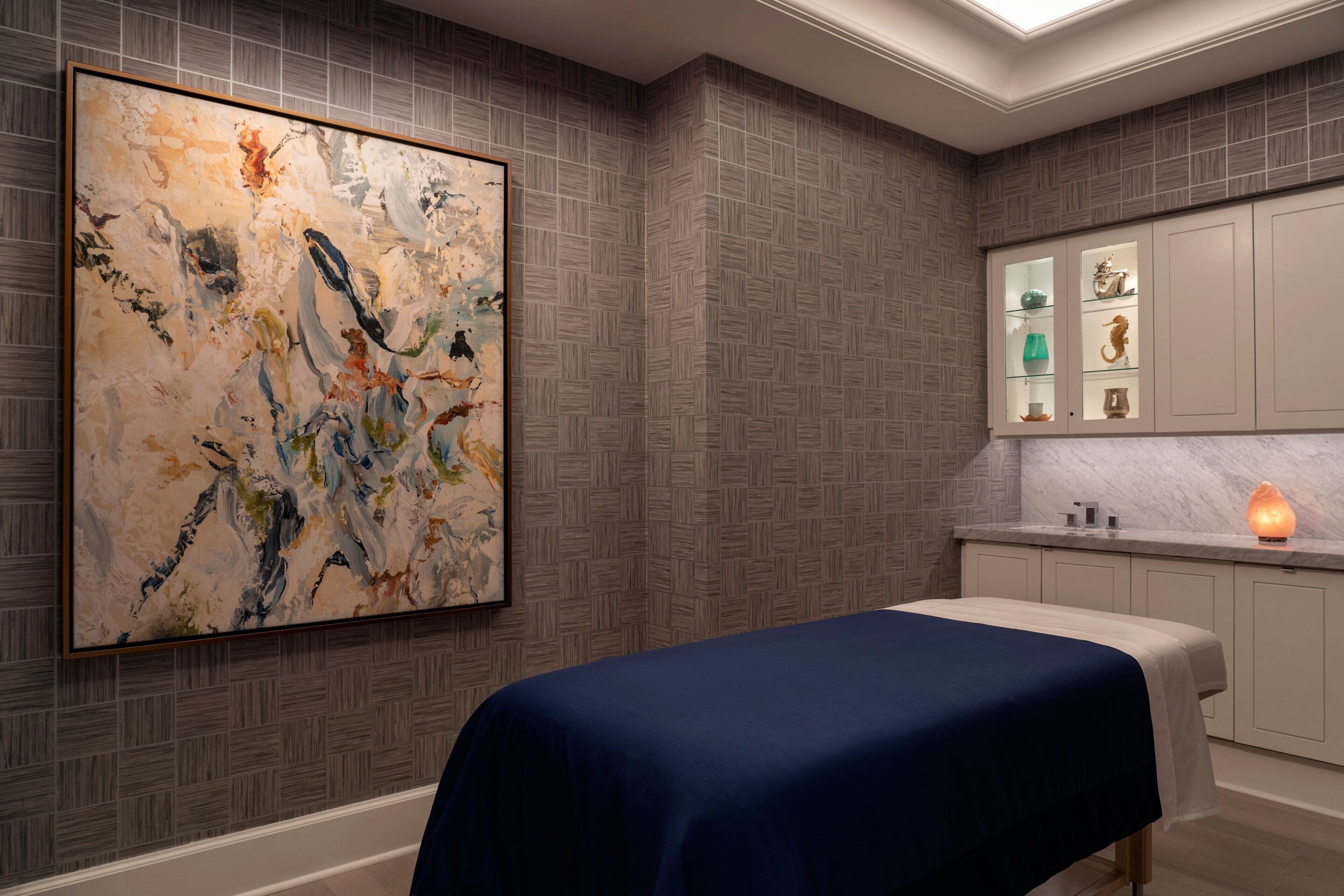 A room with a large abstract painting, a bed with a blue cover, a cabinet with glass doors, and a lit candle on the nightstand.