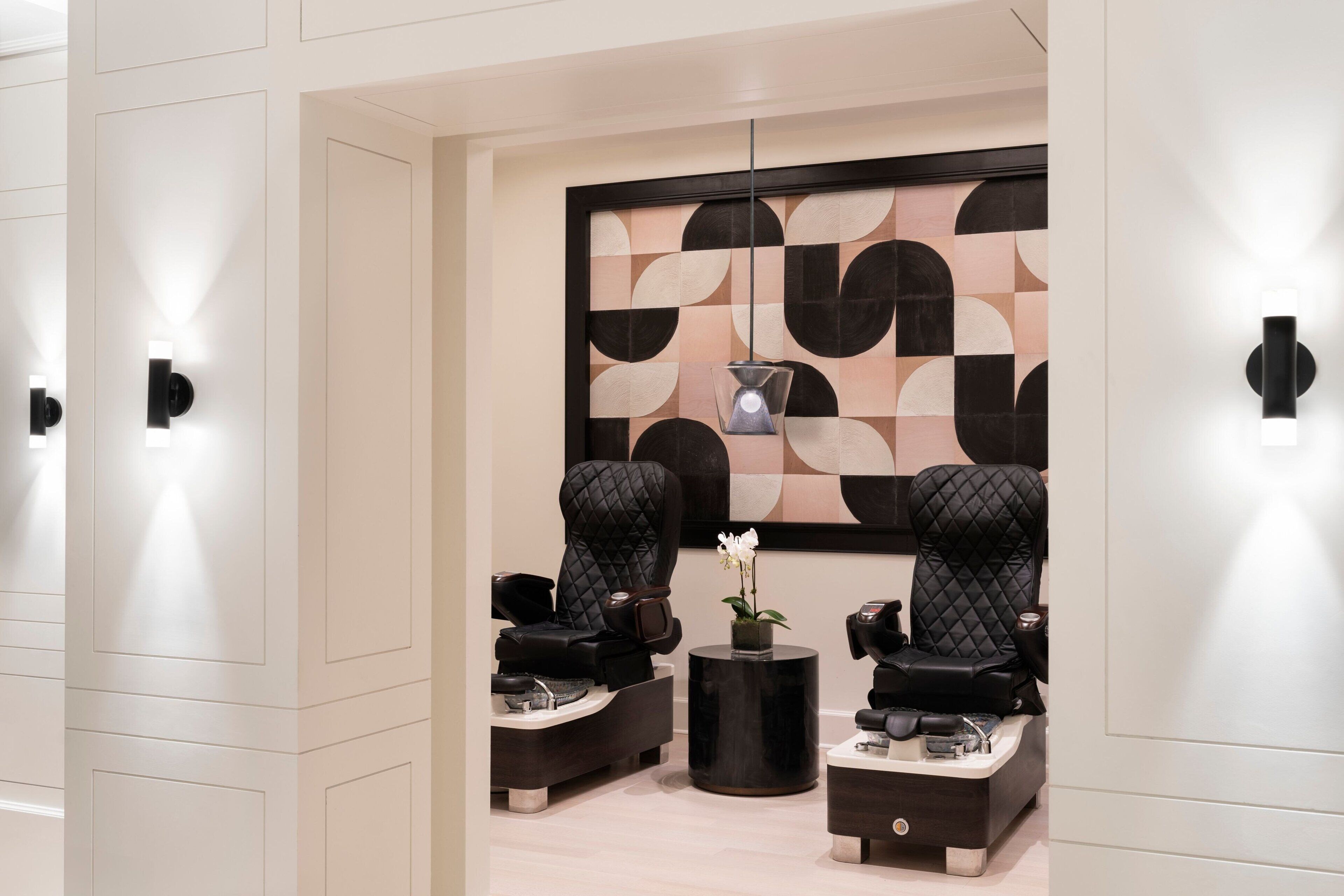 A modern spa room with two black massage chairs, a small round table with a vase of flowers, and a geometric patterned wall.
