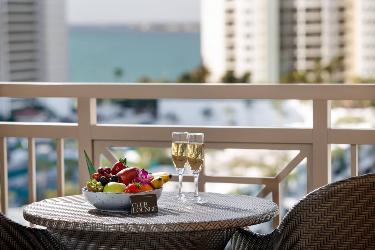 A wicker table with a fruit bowl and two glasses of champagne on a balcony overlooking a cityscape.