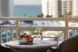 A wicker table with a fruit bowl and two glasses of champagne on a balcony overlooking a cityscape.