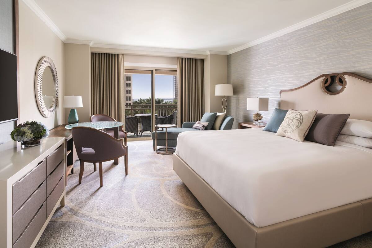 A hotel room with a large bed, a desk with a chair, a small table, and a balcony with curtains.