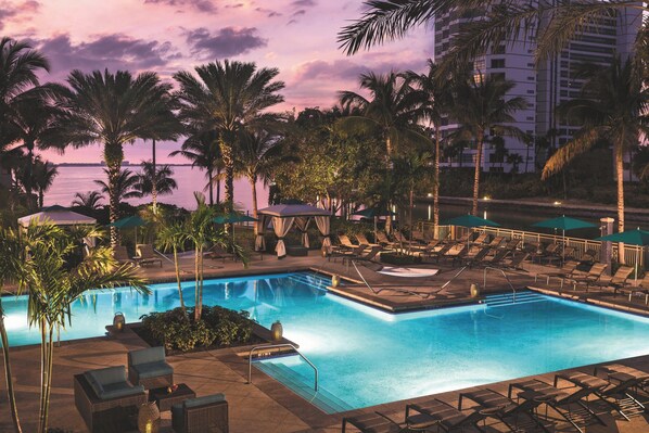 A poolside area with lounge chairs and umbrellas, surrounded by palm trees and a view of the ocean at dusk.