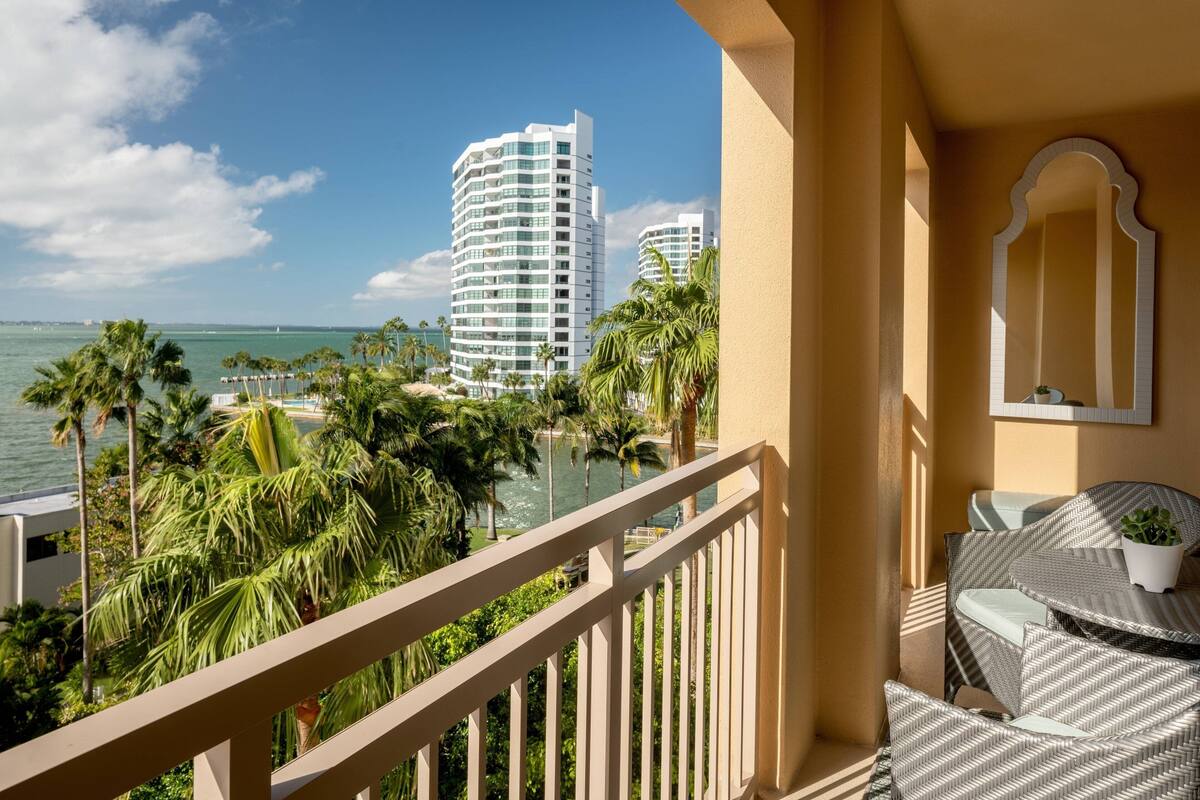 A balcony with a view of a beach and palm trees, a white railing, and a small table with a chair.
