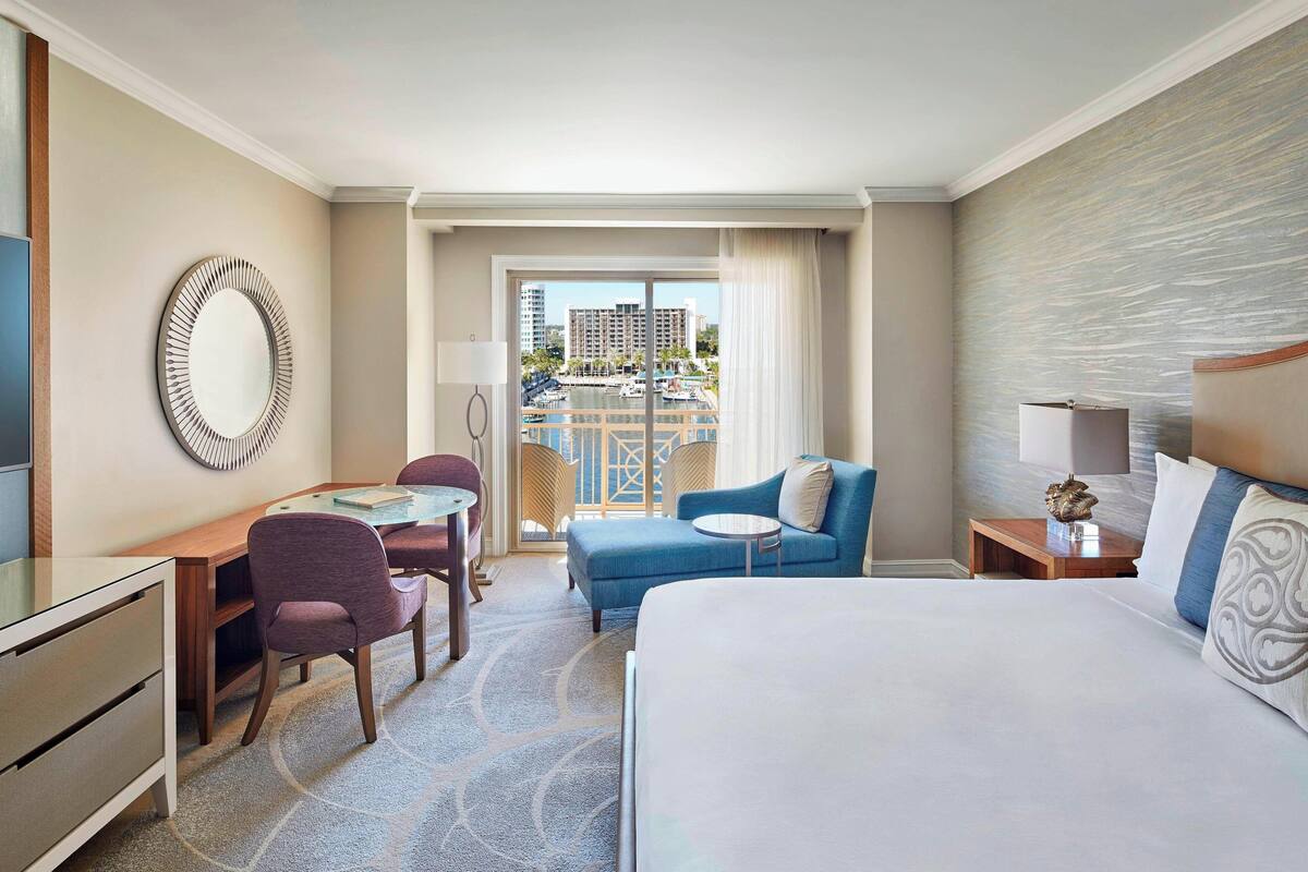 A hotel room with a bed, a desk, a chair, a small table, and a view of a marina.