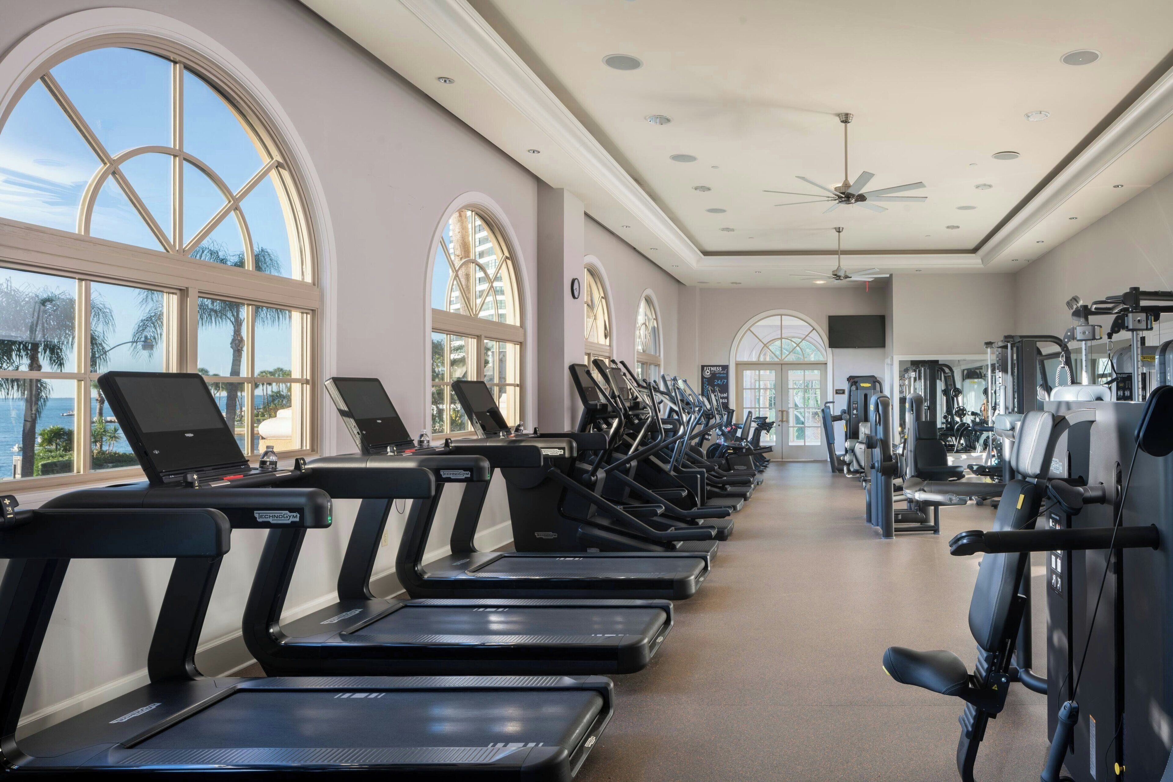 Ein Fitnessstudio mit Laufbändern, Trainingsgeräten und großen Fenstern mit Blick auf Palmen und den Ozean.
