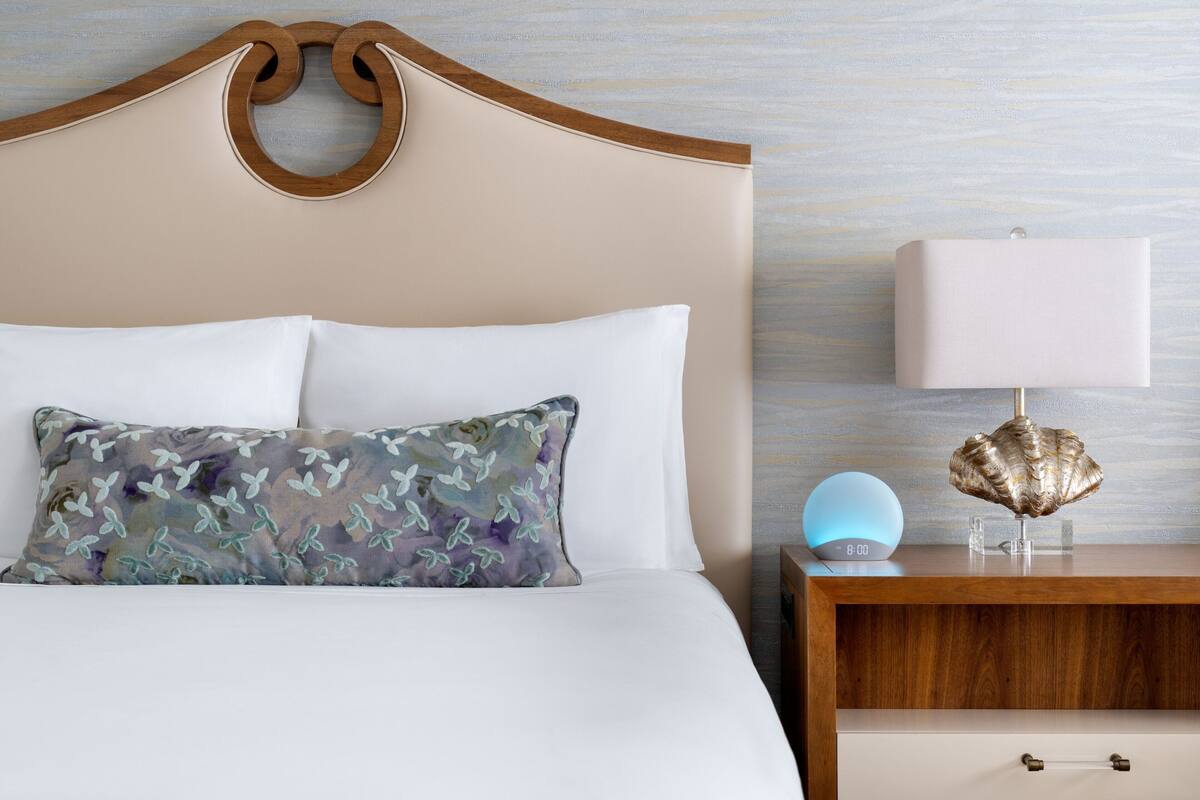 A neatly made bed with a floral pillow, a bedside lamp, and a clock on a wooden nightstand.