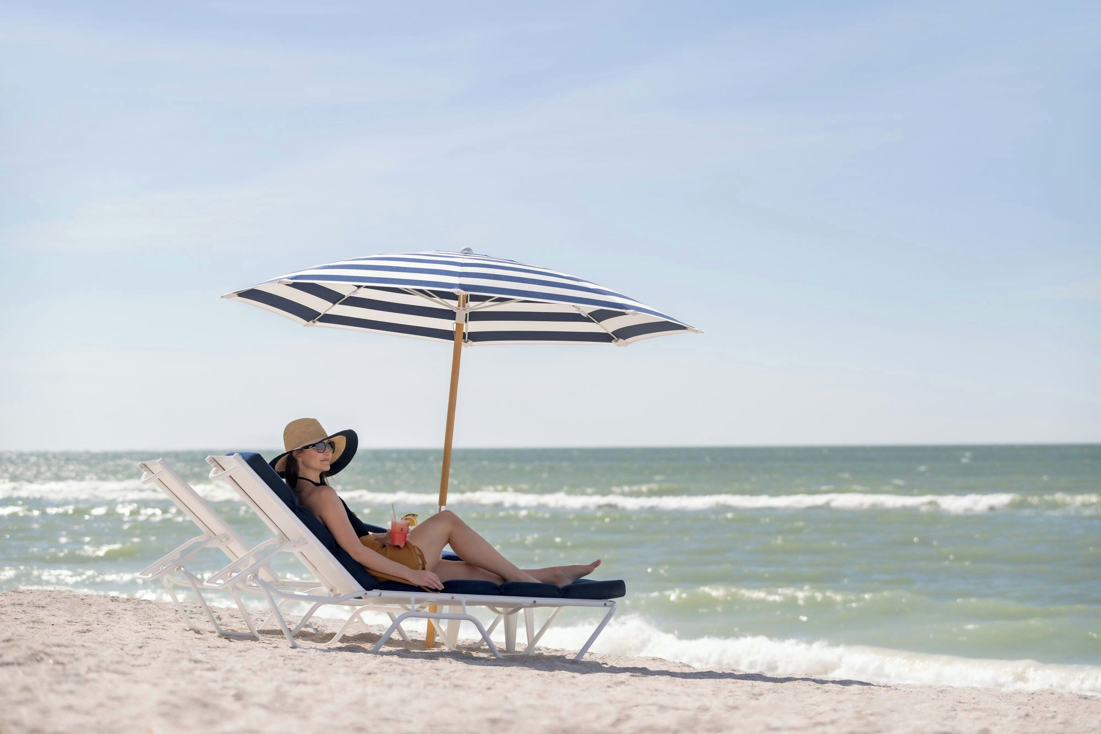 A beach scene with a person relaxing on a sun lounger under a striped umbrella.