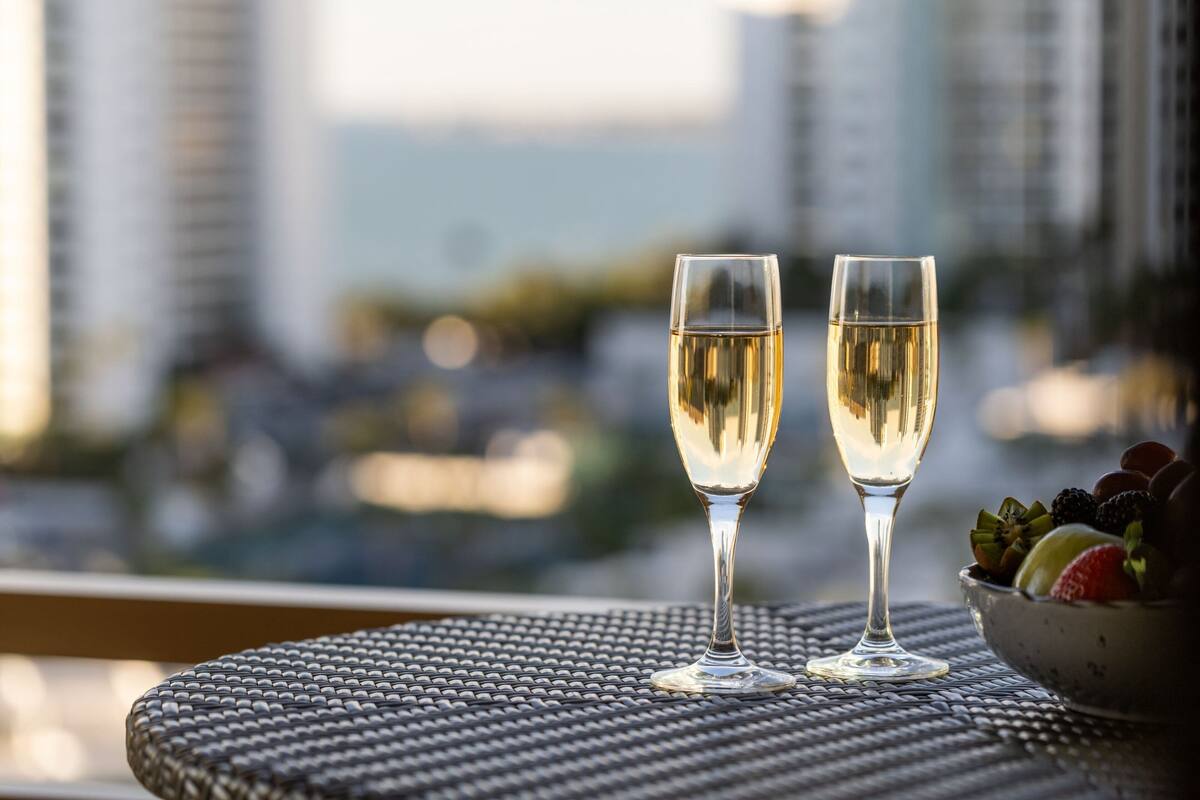 Two glasses of white wine on a round table with a woven texture, a bowl of assorted fruits, and a cityscape view in the background.