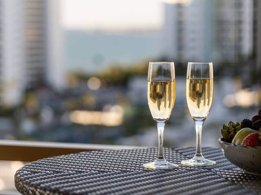 Two glasses of white wine on a round table with a woven texture, a bowl of assorted fruits, and a cityscape view in the background.