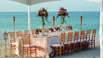 Mobiliario para comidas frente al mar, con una mesa dispuesta para varios comensales, sillas y centros de mesa con flores.