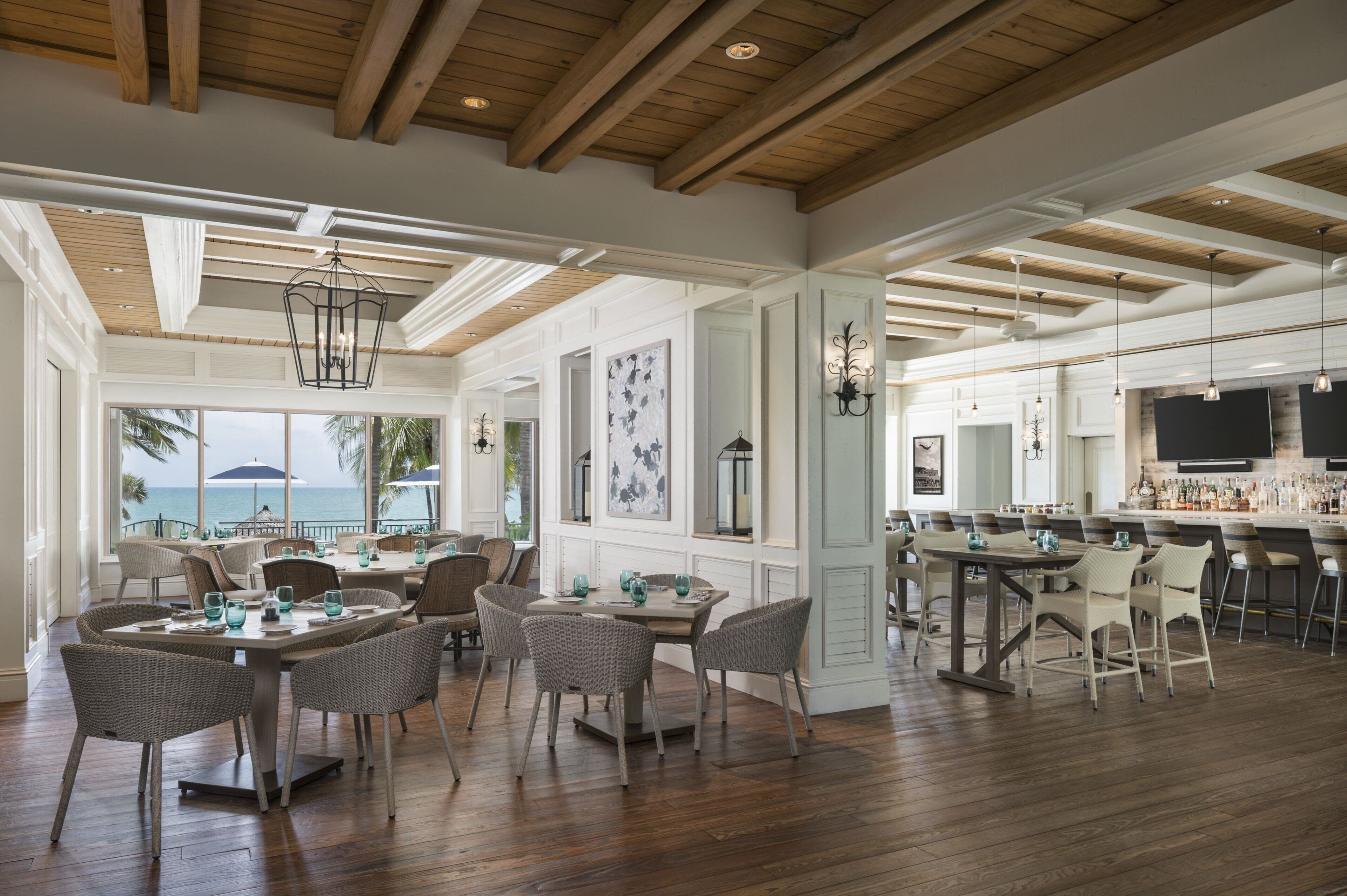 Un vaste espace dédié à la salle à manger, doté de hauts plafonds et de poutres en bois, comprenant plusieurs tables et chaises, un bar et de grandes fenêtres offrant une vue sur l’océan.