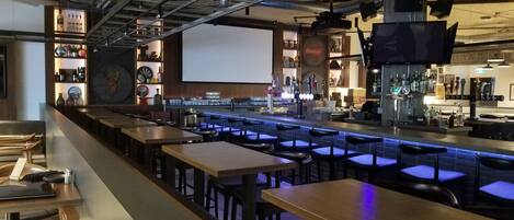 A bar area with multiple seating tables and stools, a bar counter with a variety of bottles and glasses, and a large screen on the wall.