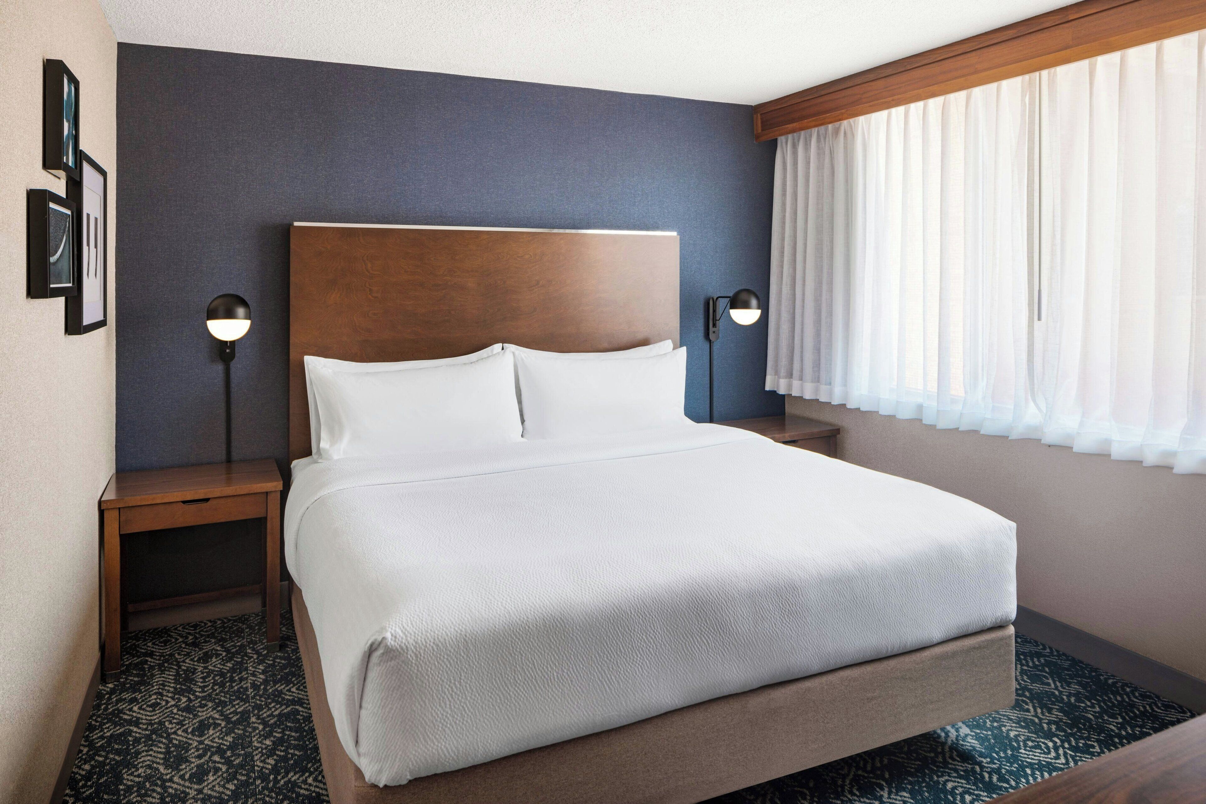 A hotel room with a large bed, two bedside tables, a headboard, and a window with curtains.