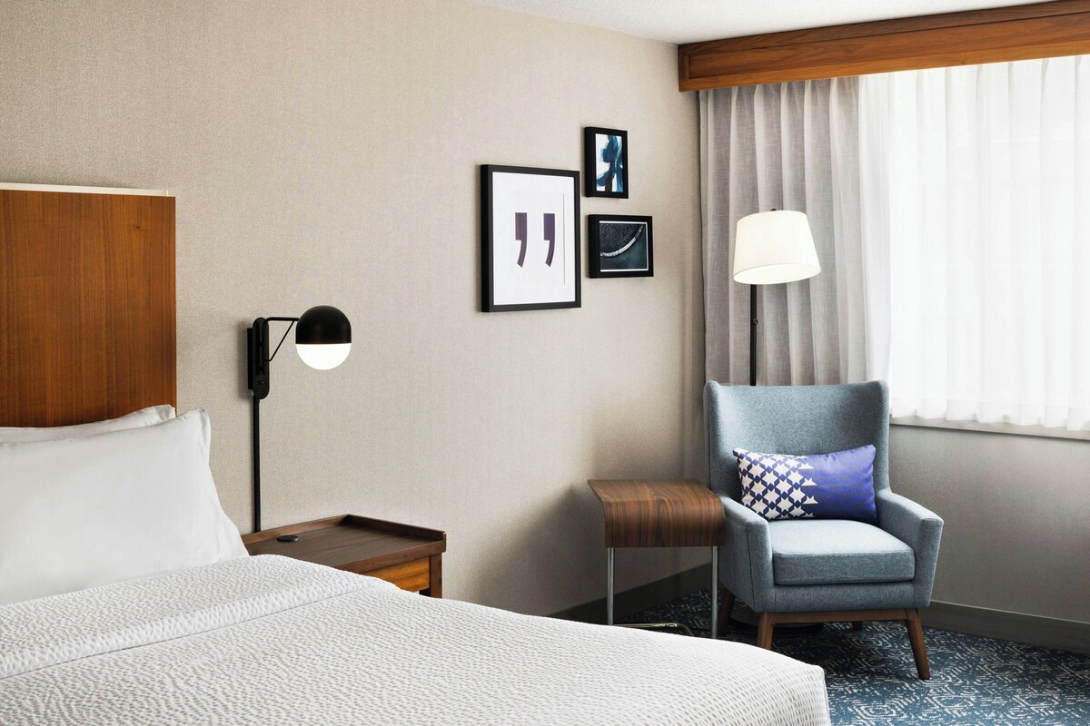 A hotel room with a bed, a chair, a nightstand, a lamp, and framed pictures on the wall.