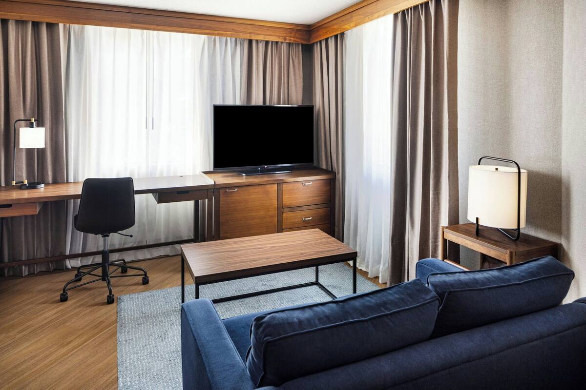 A hotel room with a flat-screen TV, wooden desk, chair, sofa, and bedside table with lamps.