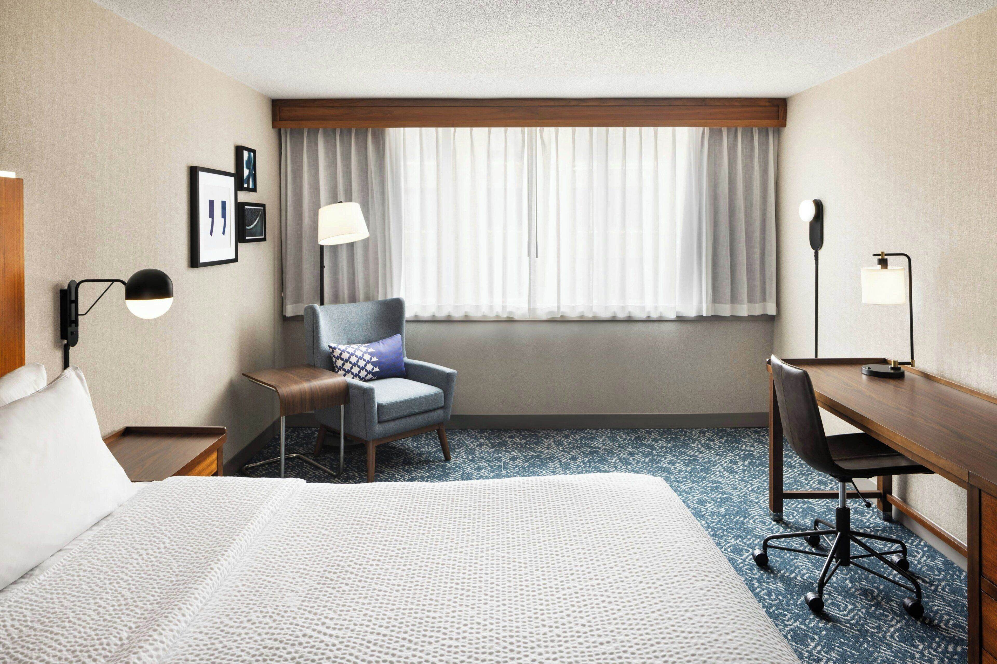 A hotel room with a large bed, a desk, a chair, a desk lamp, a floor lamp, and a window with curtains.