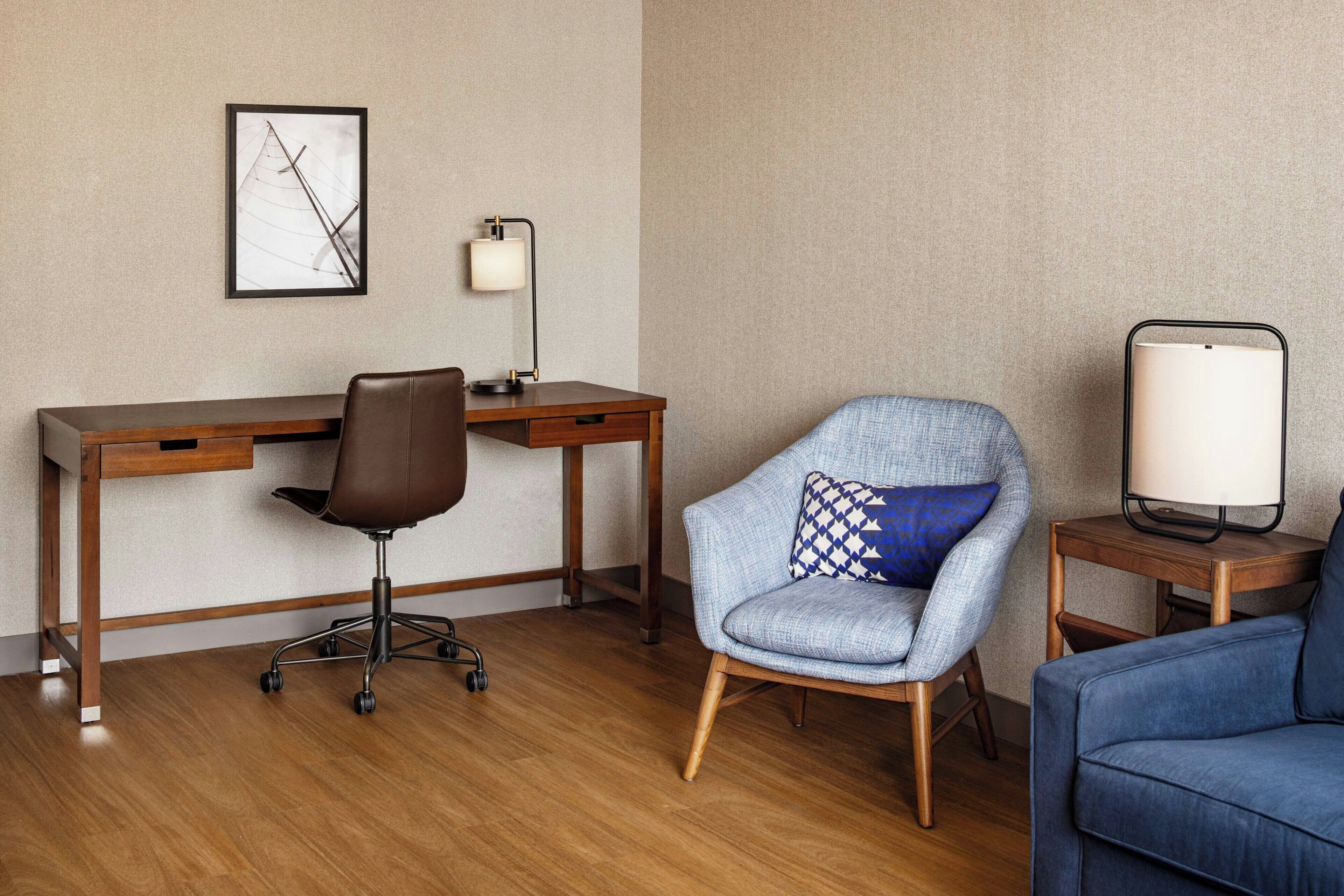 Et hotelværelse med et træskrivebord, en drejestol, en blå lænestol med et mønstret pude, en blå sofa og et natbord med en lampe.